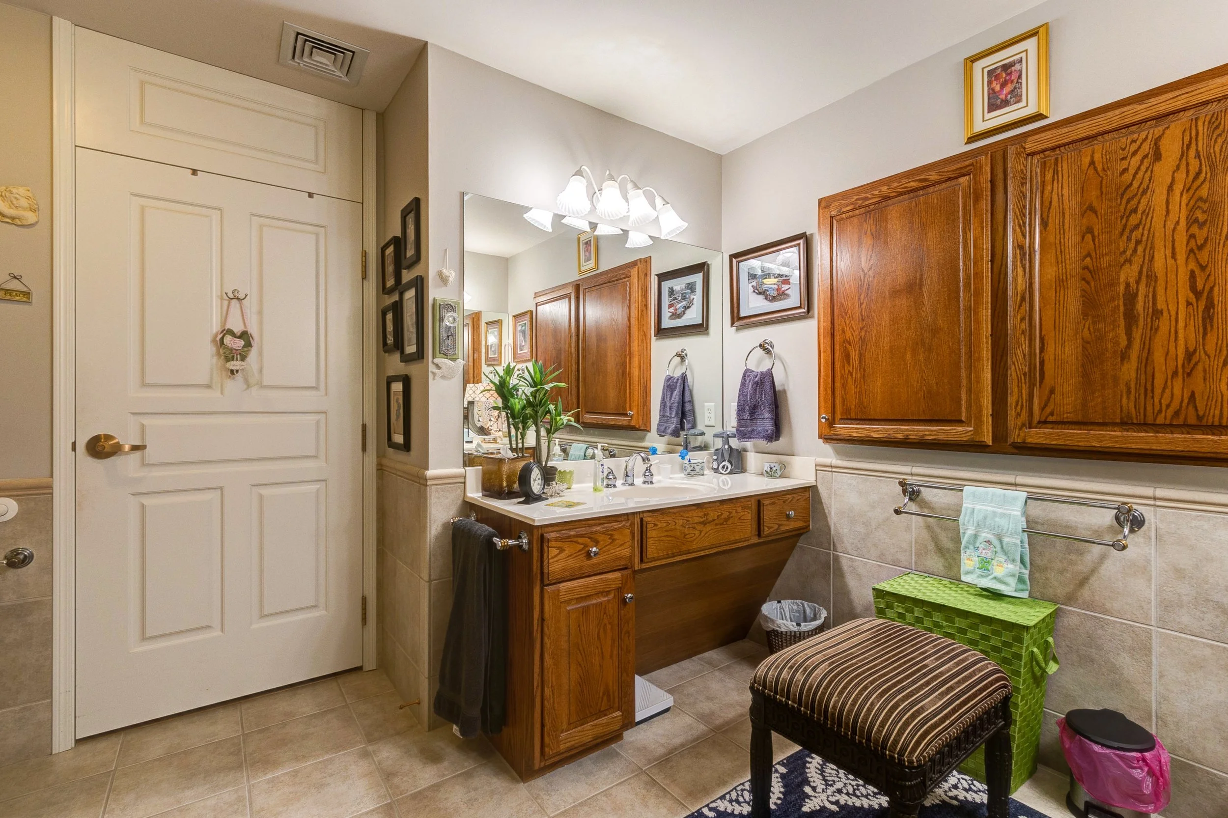 Bathroom with wooden cabinets, a large mirror, hanging towels, framed pictures on the wall, a plant, and various toiletries.