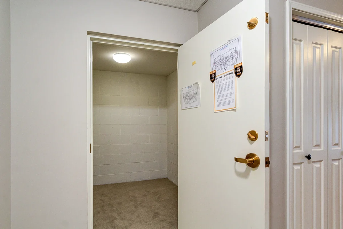 Open white door revealing a small, empty room with beige carpet and white cinder block walls, illuminated by a ceiling light.