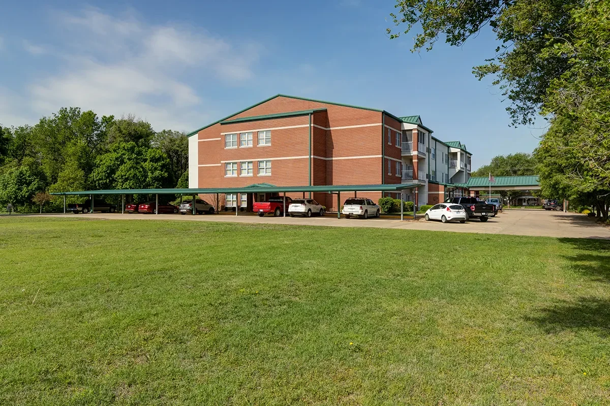 A three-story brick apartment building with a green metal roof, surrounded by a parking lot with several cars, and green trees on both sides, under a partly cloudy sky.