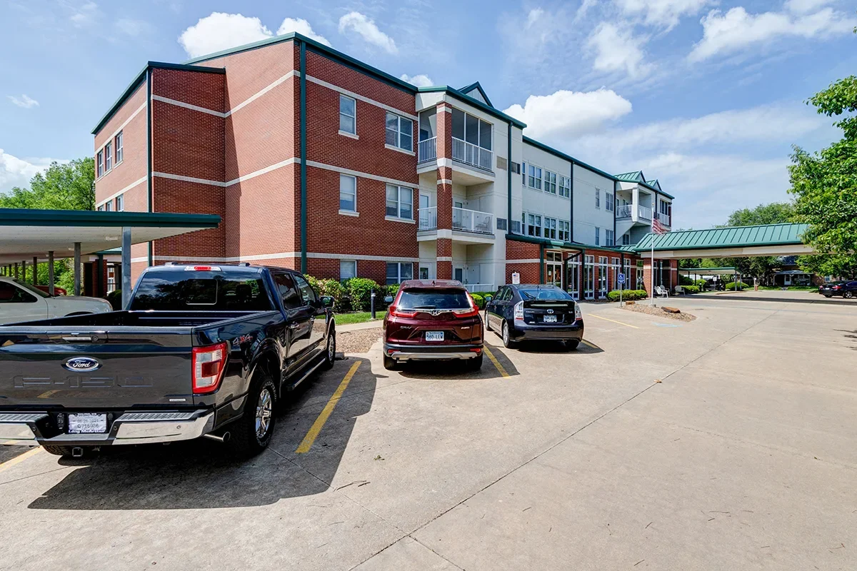 Parking lot in front of a multi-story residential apartment complex with several parked cars and a covered walkway, under a partly cloudy sky.