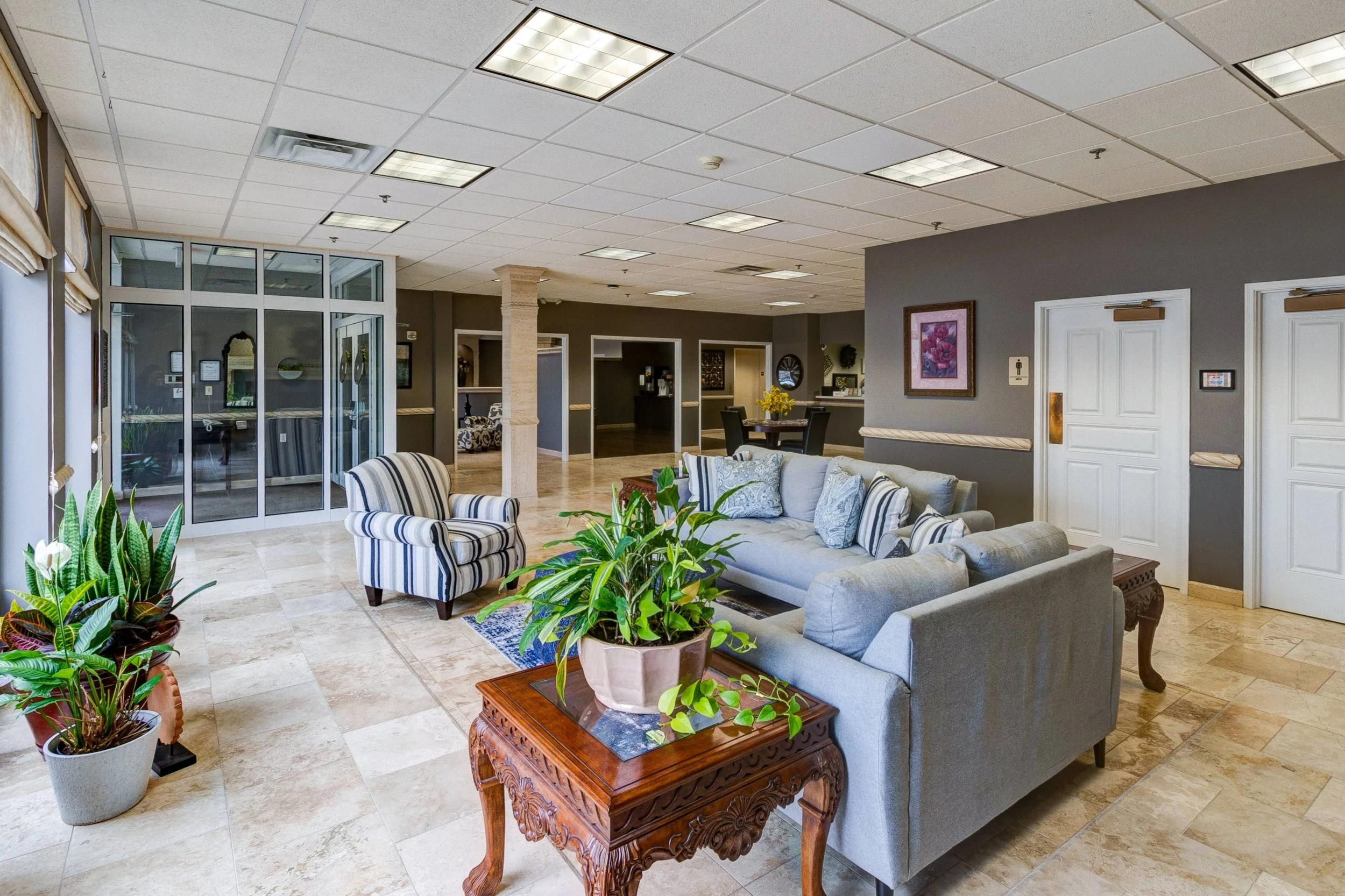 Interior view of a hotel lobby with seating area including a gray sofa with decorative pillows, an armchair with blue and white stripes, and potted plants. The space has tiled floors, a drop ceiling with fluorescent lighting, and a background recepti