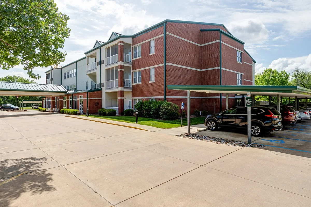 A multi-story brick and siding apartment complex with a parking lot and covered parking spaces, surrounded by trees and greenery, under a partly cloudy sky.