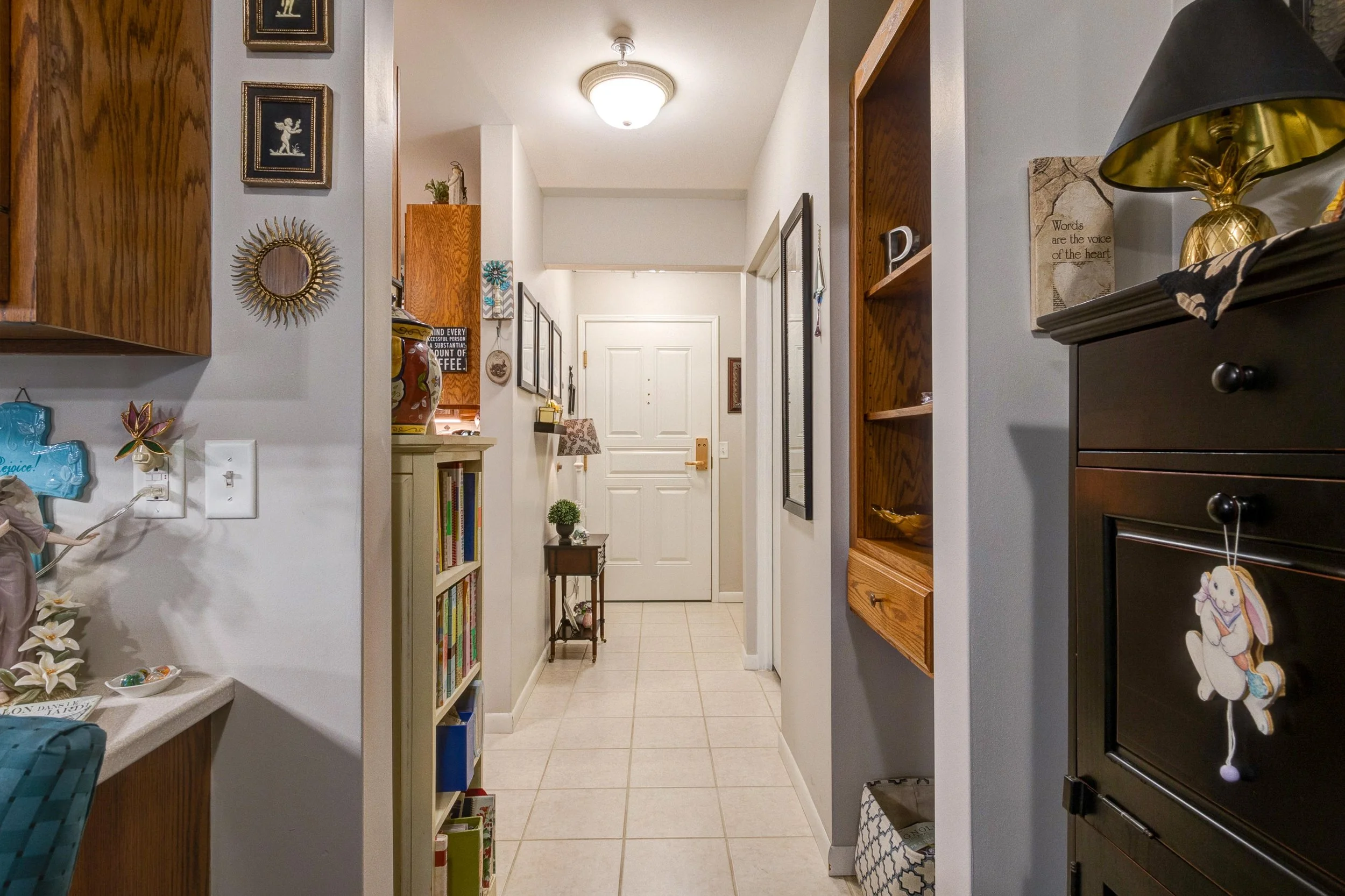 View of a hallway in a home with white walls and tiled floor, decorated with wall art, bookshelves, and a small table with a potted plant, leading to a white front door.