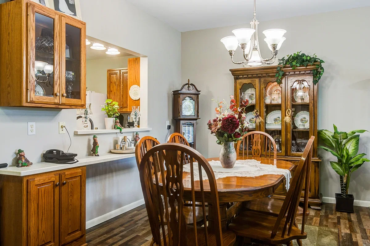 A dining room with a wooden table and six matching chairs, a glass-front hutch filled with dishes, a grandfather clock, a chandelier above the table, and various decorative plants and items.