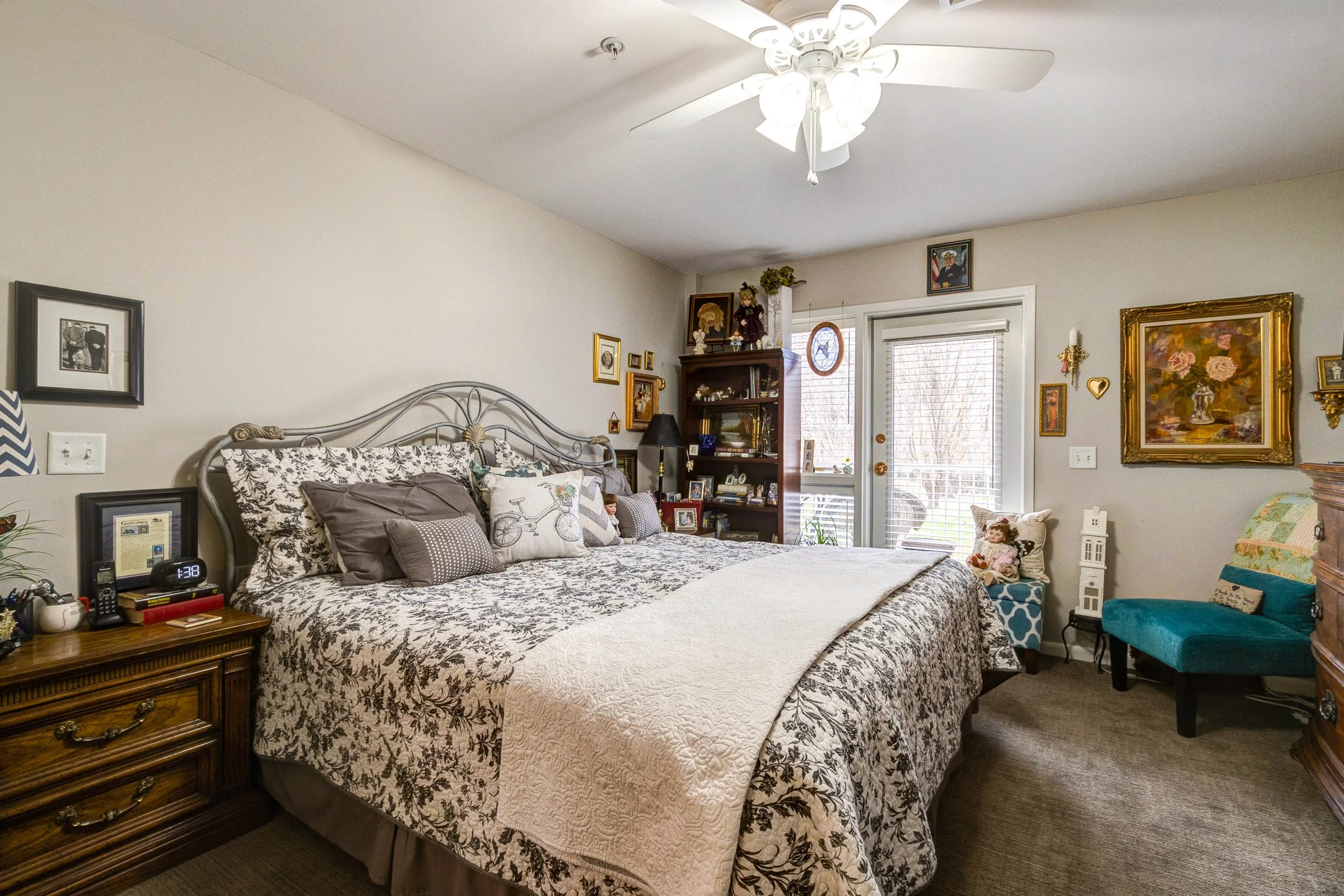 A bedroom with a large bed with black and white floral bedding and multiple pillows. To the left is a wooden nightstand with a digital clock, picture frame, and other items. There is a ceiling fan above. To the right are a teal upholstered chair, a s