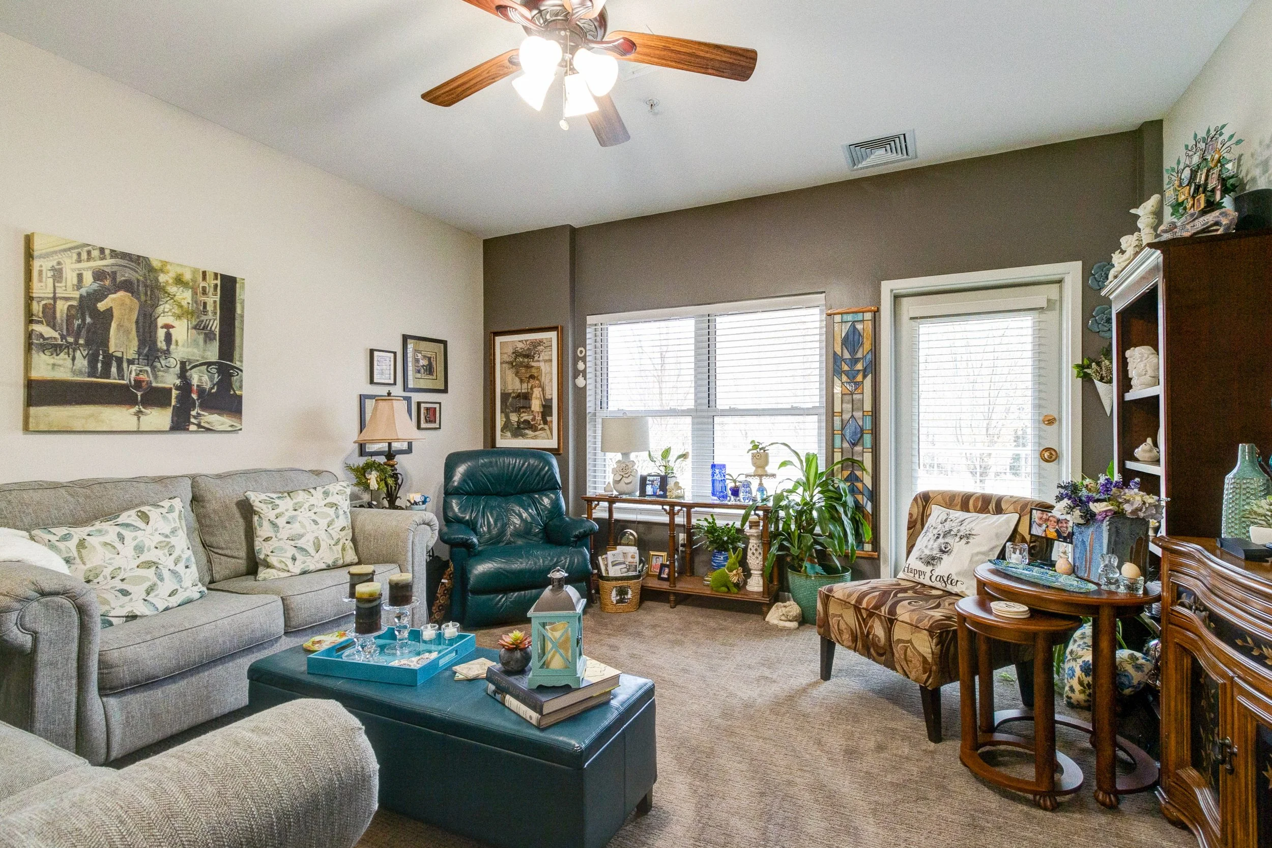 Living room with a beige couch, a green leather chair, and a patterned side chair. Decor includes paintings, plants, and small decorative items on tables and shelves. Large window with blinds and a door with blinds lead outside. Ceiling fan with ligh