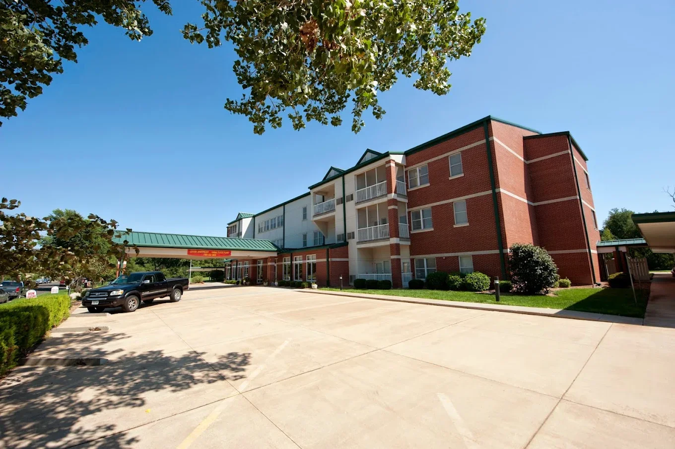 A three-story residential building with a brick exterior and white balconies, situated under a clear blue sky, with a parking lot and some cars in front, and surrounded by greenery.