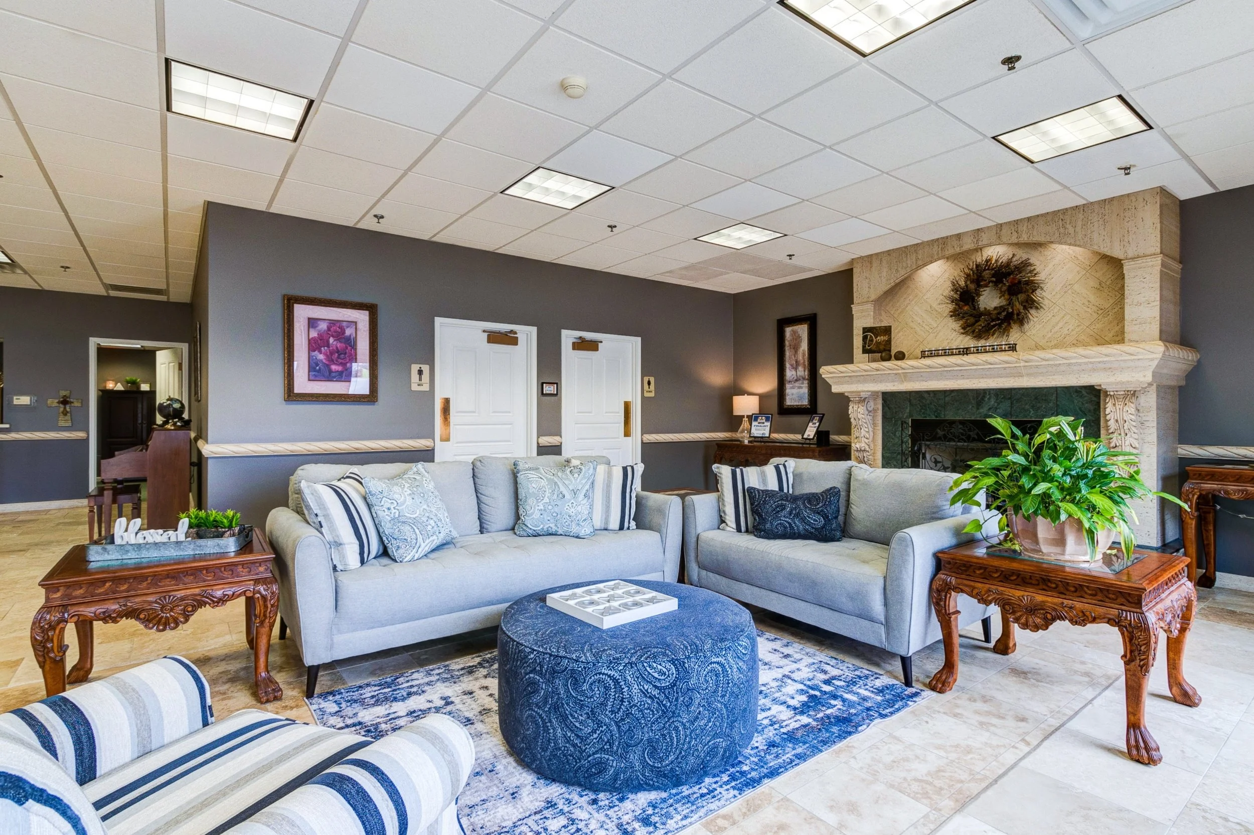 A cozy hotel lounge with two beige sofas, striped and patterned throw pillows, a blue patterned ottoman, and wooden side tables with plants, artwork on the wall, and a fireplace with a decorative wreath above.