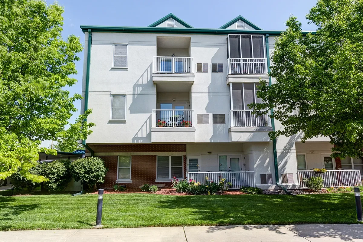 Exterior view of a four-story apartment building with white walls, green roof, and balconies. Green trees and a well-kept lawn are in the foreground.