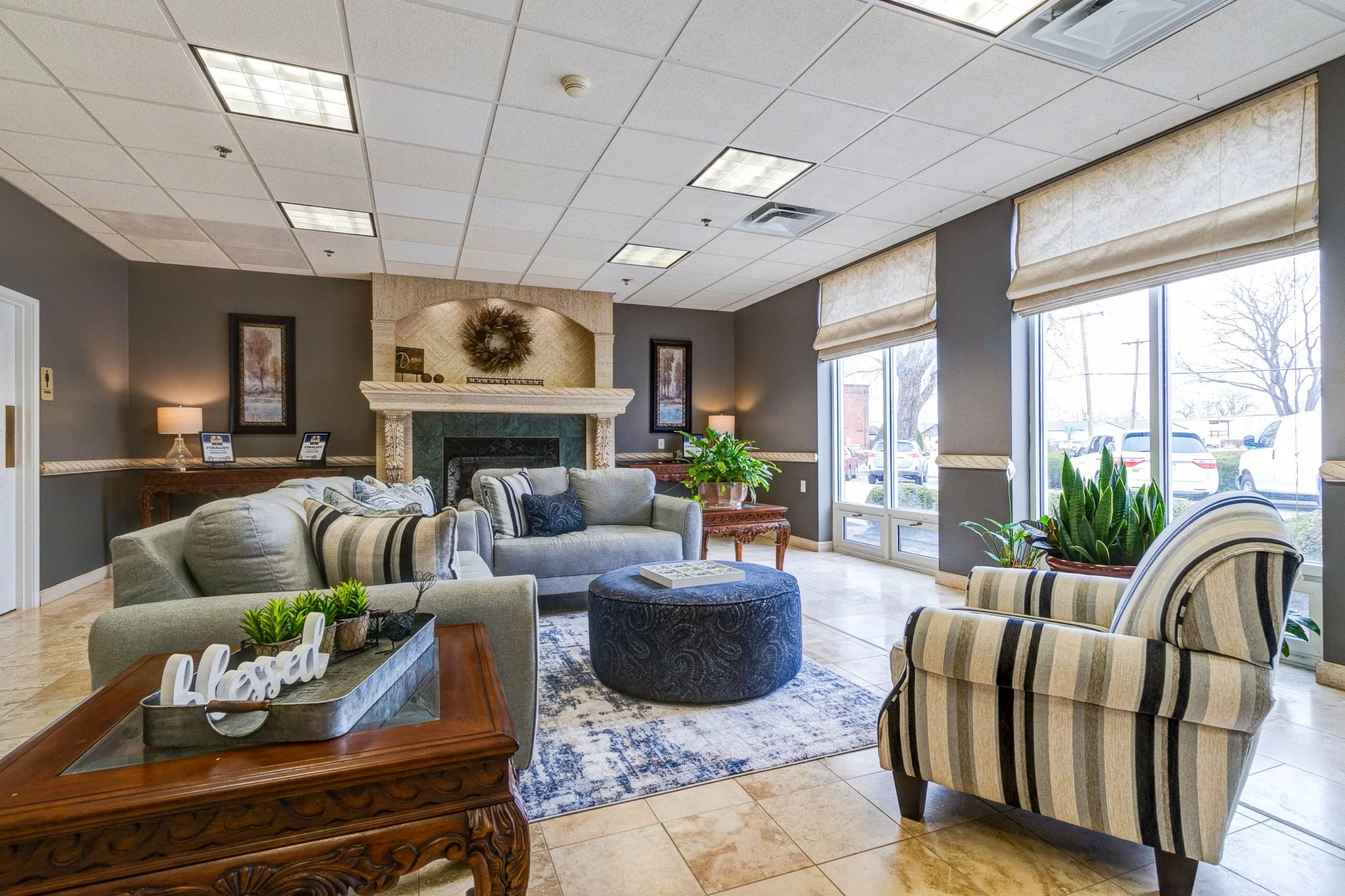 A cozy lobby with gray walls, a fireplace, and wooden accents. Comfortable sofas, armchairs, and a round ottoman fill the space, accented by houseplants and decorative pillows. Large windows provide natural light and a view outside.