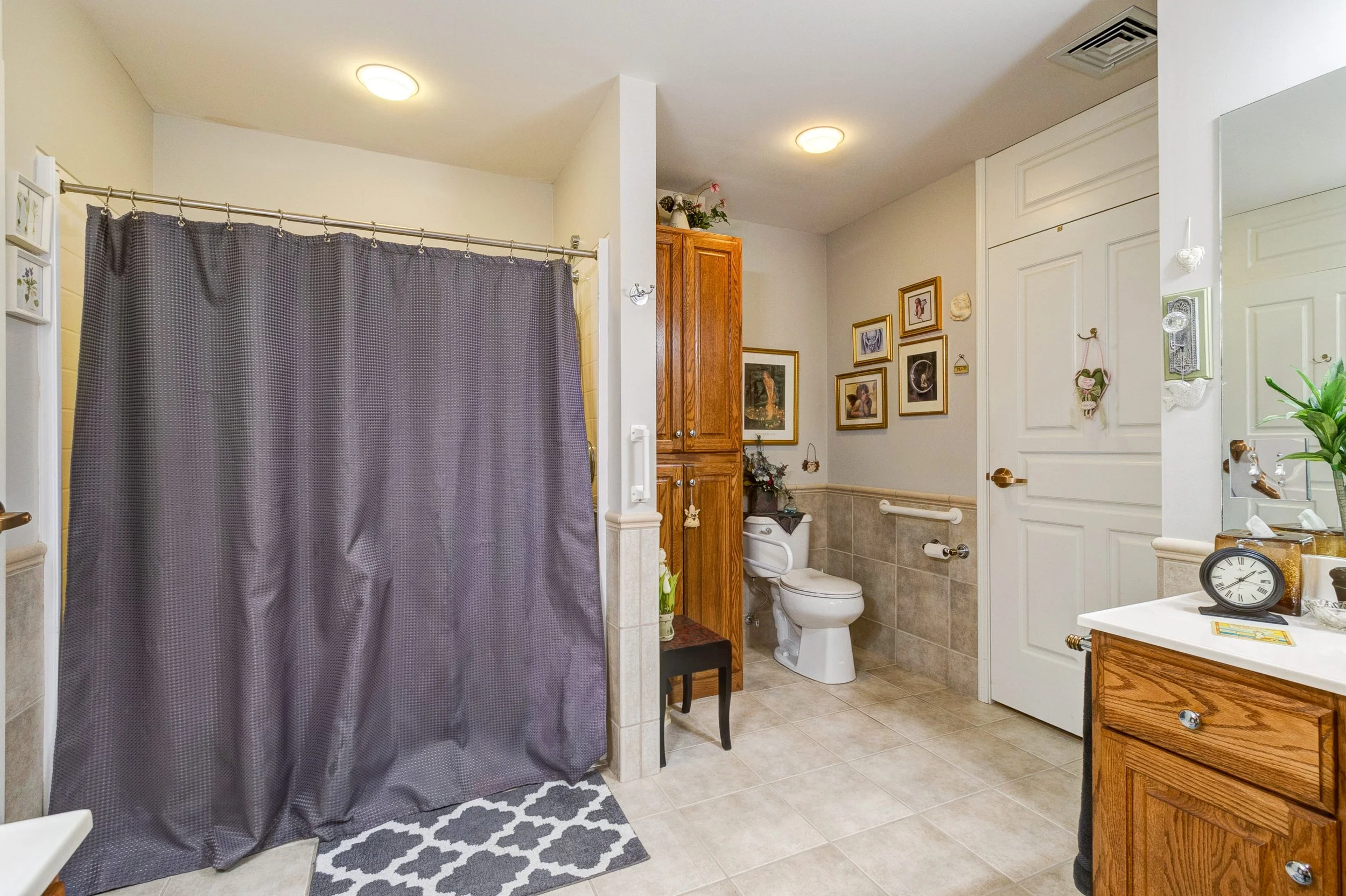 Bathroom with a grey shower curtain, beige tiled floor, wooden cabinet, decorative wall art, toilet, and sink with a clock and plants.