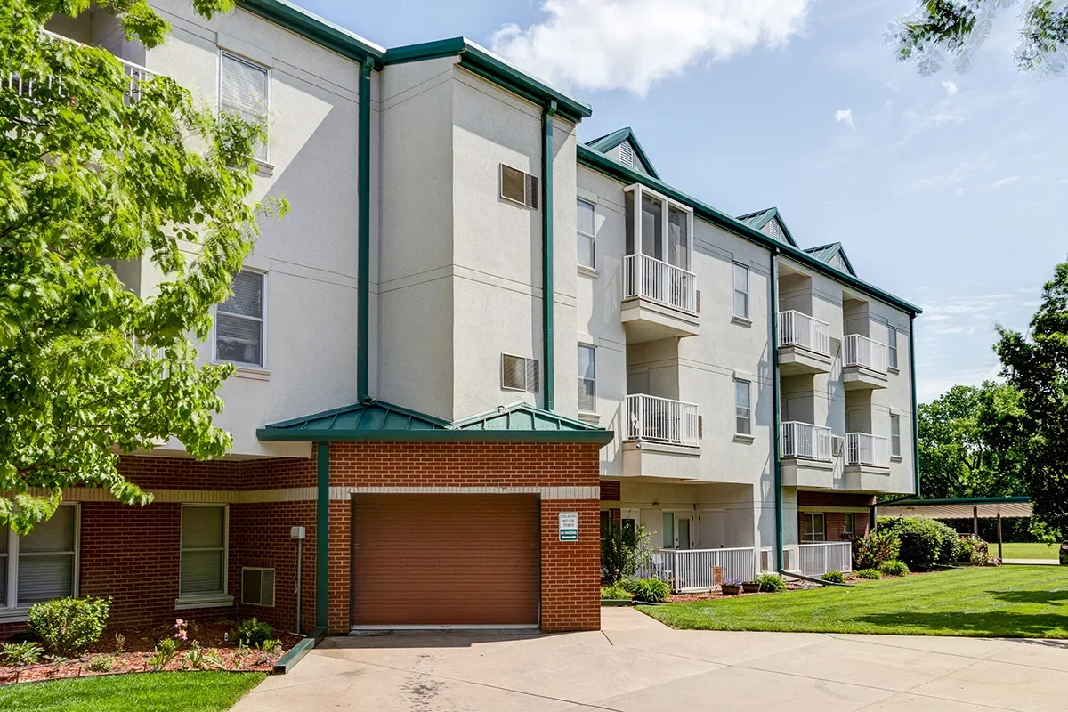 Exterior view of a modern four-story apartment building with balconies, a green roof, and a brick garage door under a partly cloudy sky, surrounded by green trees and a well-maintained lawn.