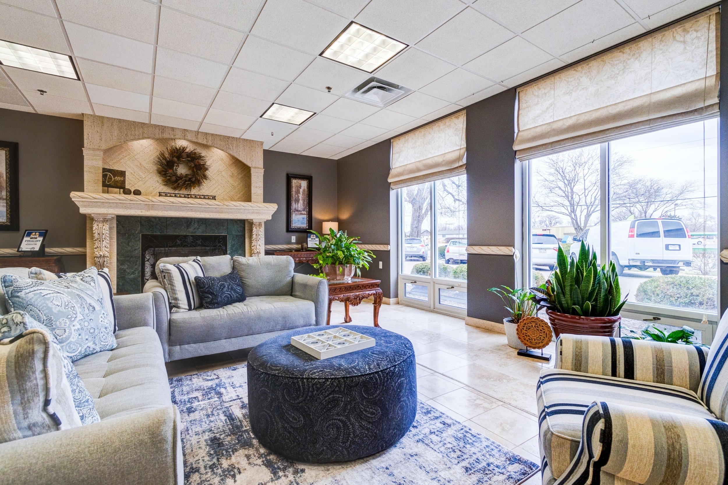 A cozy waiting area with multiple sofas and chairs, a fireplace, large windows with beige blinds, green plants, and decorative items.