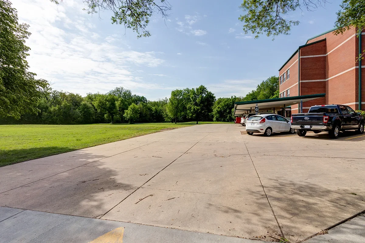 Empty parking lot with a few parked cars near a brick building and green trees under a partly cloudy sky.