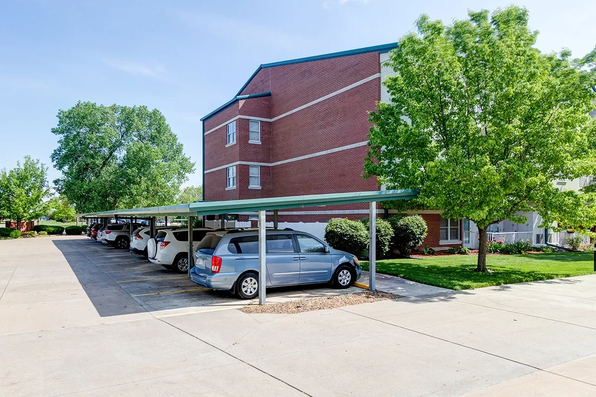 Carport with parked cars next to an apartment building, trees, and a clean parking lot.