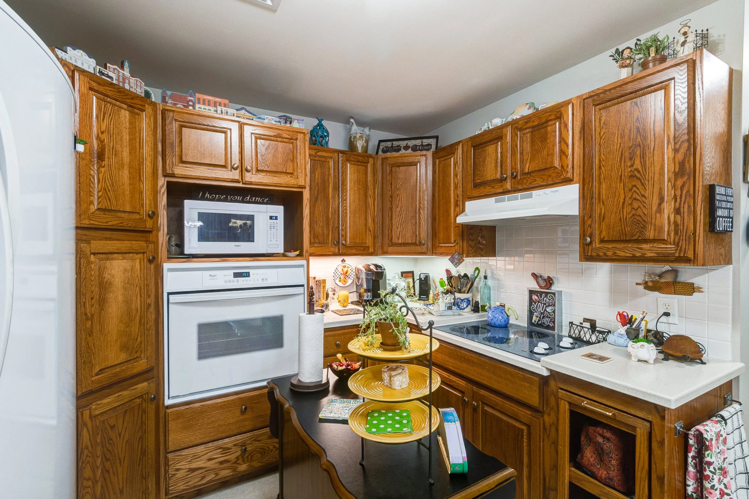A cozy kitchen with wooden cabinets, a white oven, microwave, and various kitchen items on the counters. A small black table with a yellow tiered tray and a potted plant is in the center of the kitchen.