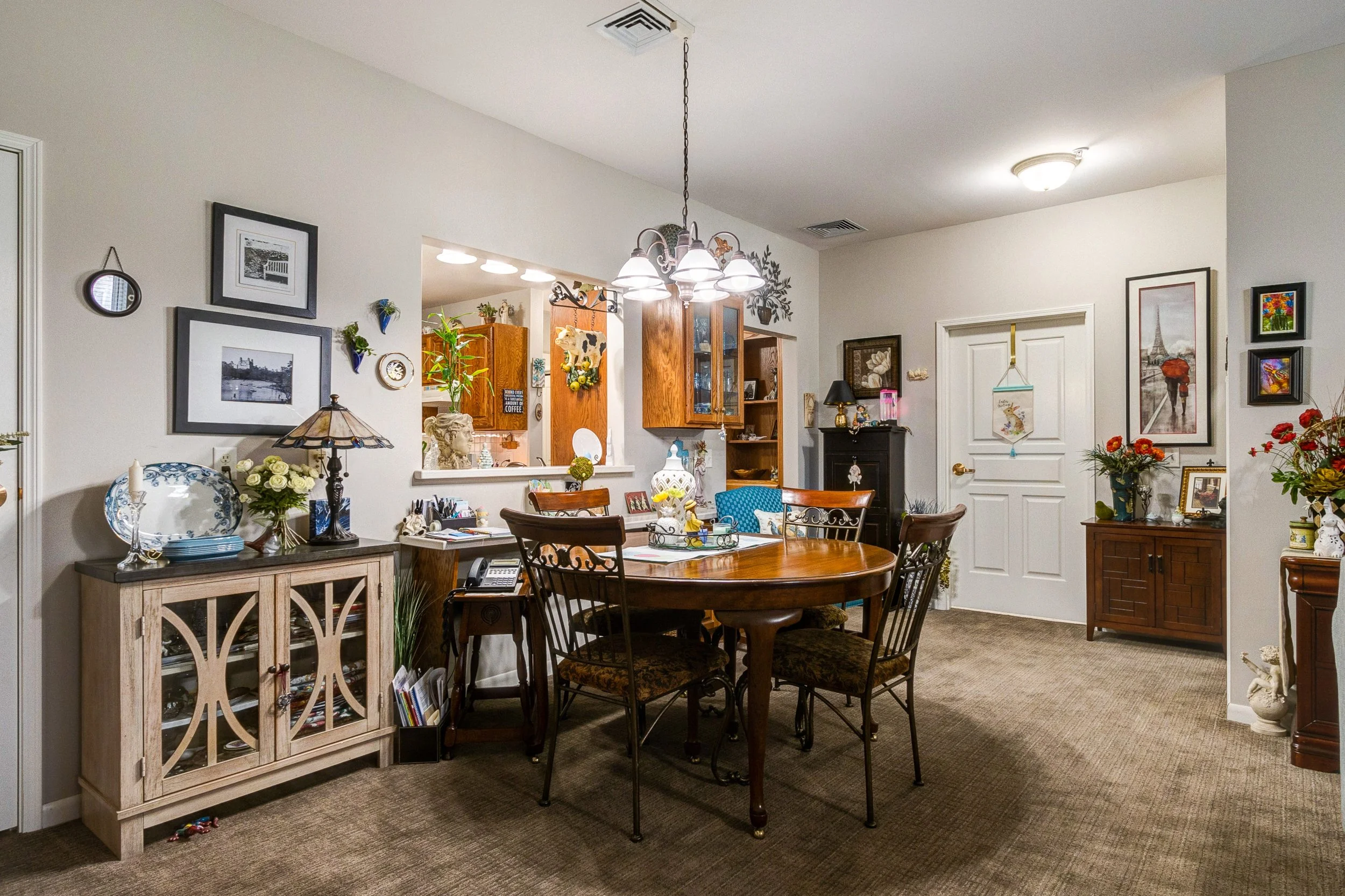 A dining room with a wooden table surrounded by six chairs, decorated with lamps, flowers, and ornaments. The room has framed pictures, a cabinet, and a view into a kitchen area.