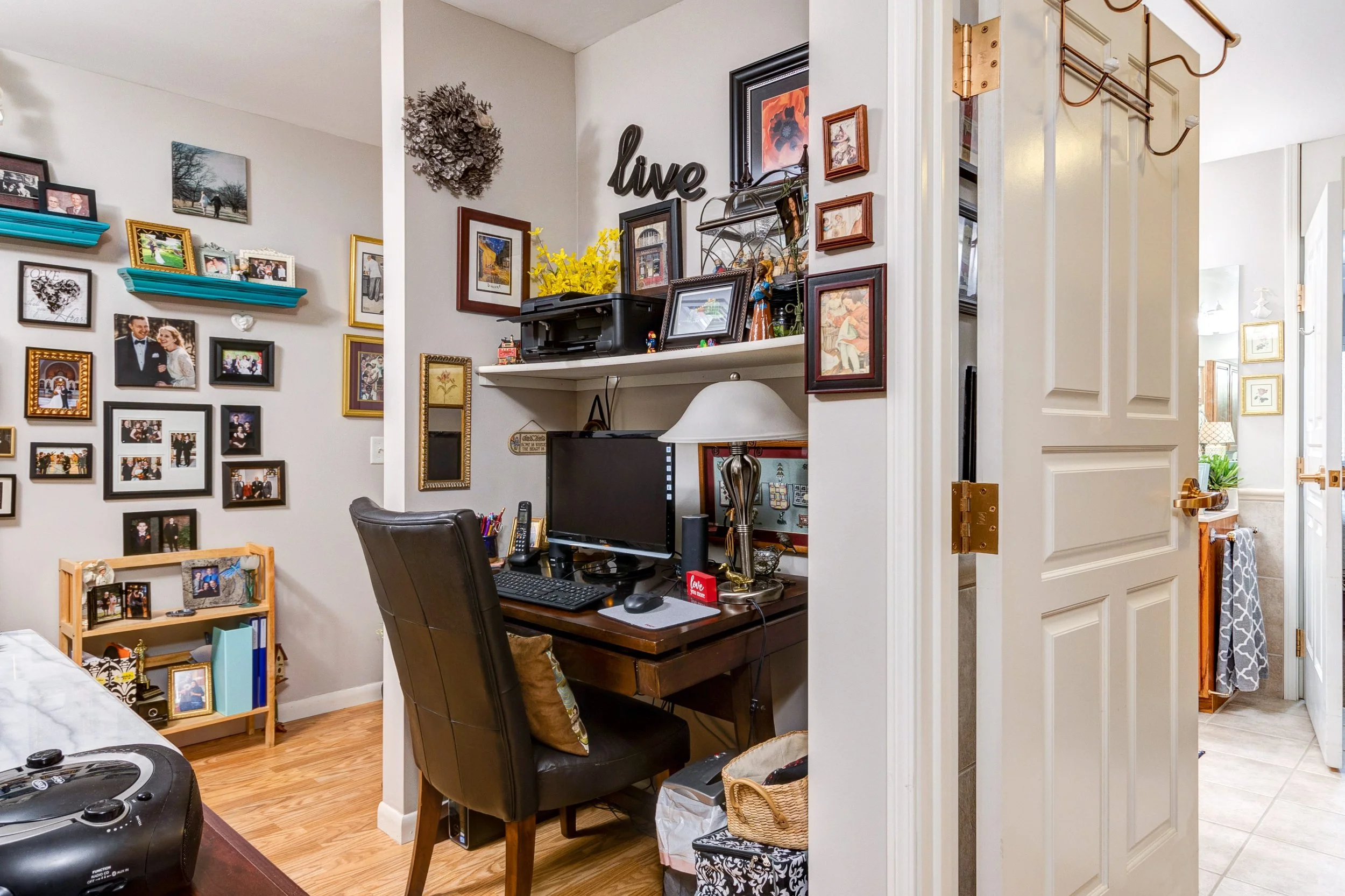 A cozy corner observed from a living room, featuring a black desk with a computer monitor, a lamp, and various decorations. The wall behind the desk is decorated with family photos, framed pictures, and artwork, including a "live" sign. To the right,
