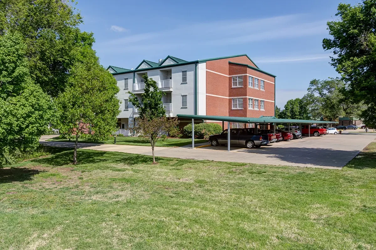Apartment building with covered parking lot in front, surrounded by green trees and lawn under a clear blue sky.