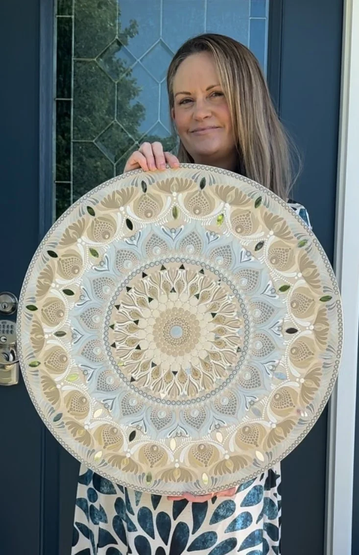 Woman holding a large decorative circular artwork with intricate gold, white, and black patterns, standing in front of a blue door with glass panels.