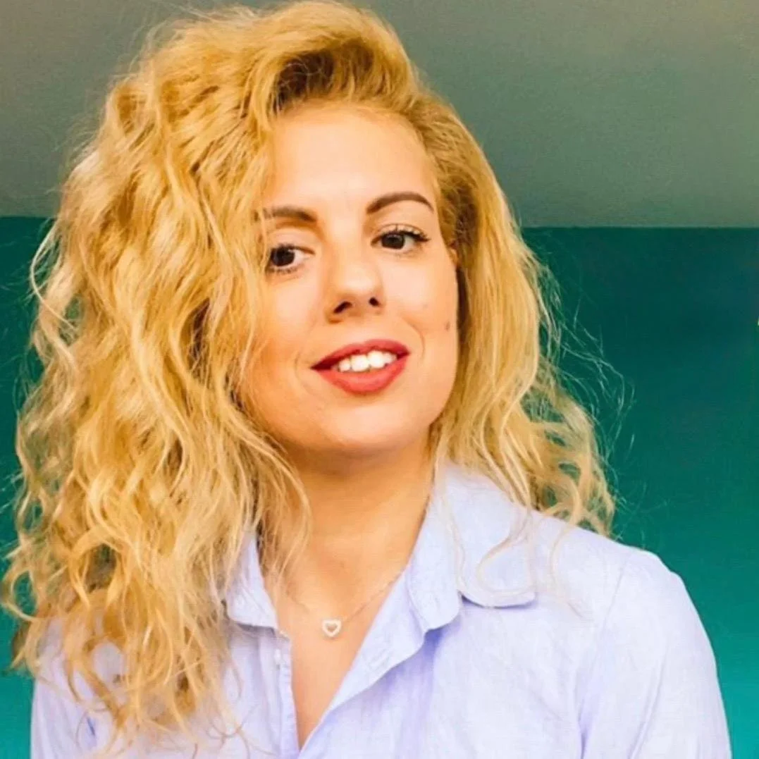 A woman with long, curly blonde hair, light skin, wearing a light blue collared shirt, and a silver heart-shaped necklace, smiling slightly against a teal background.