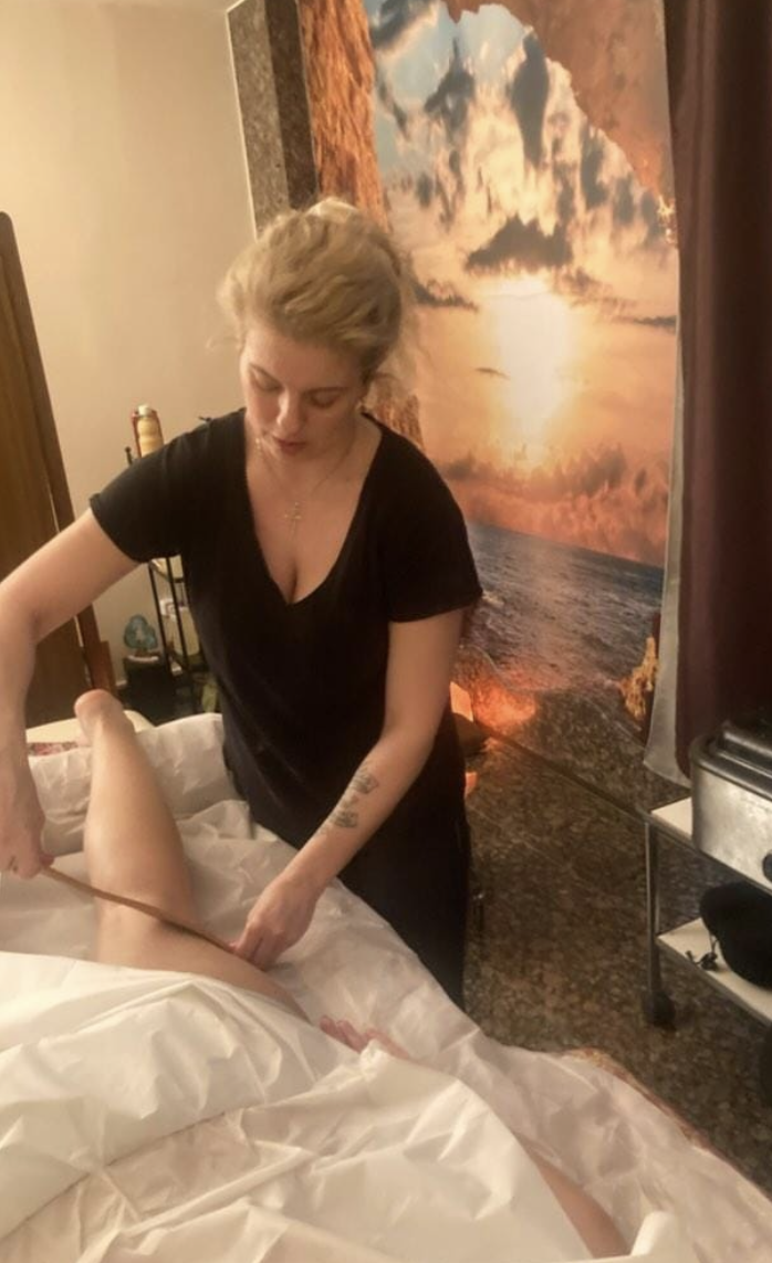 A woman in black attire giving a massage to a person lying in a bed, with a scenic sunset mural of clouds and ocean on the wall behind them.