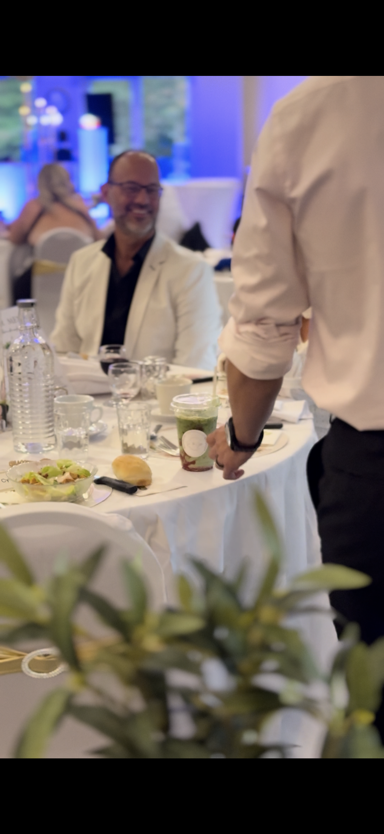 Guest at a luxury Hudson, QC wedding reception enjoying Café Apéro catering. A man in a white blazer smiles at a formal dinner table while a server delivers a signature iced matcha latte in the foreground, showcasing professional wedding beverage