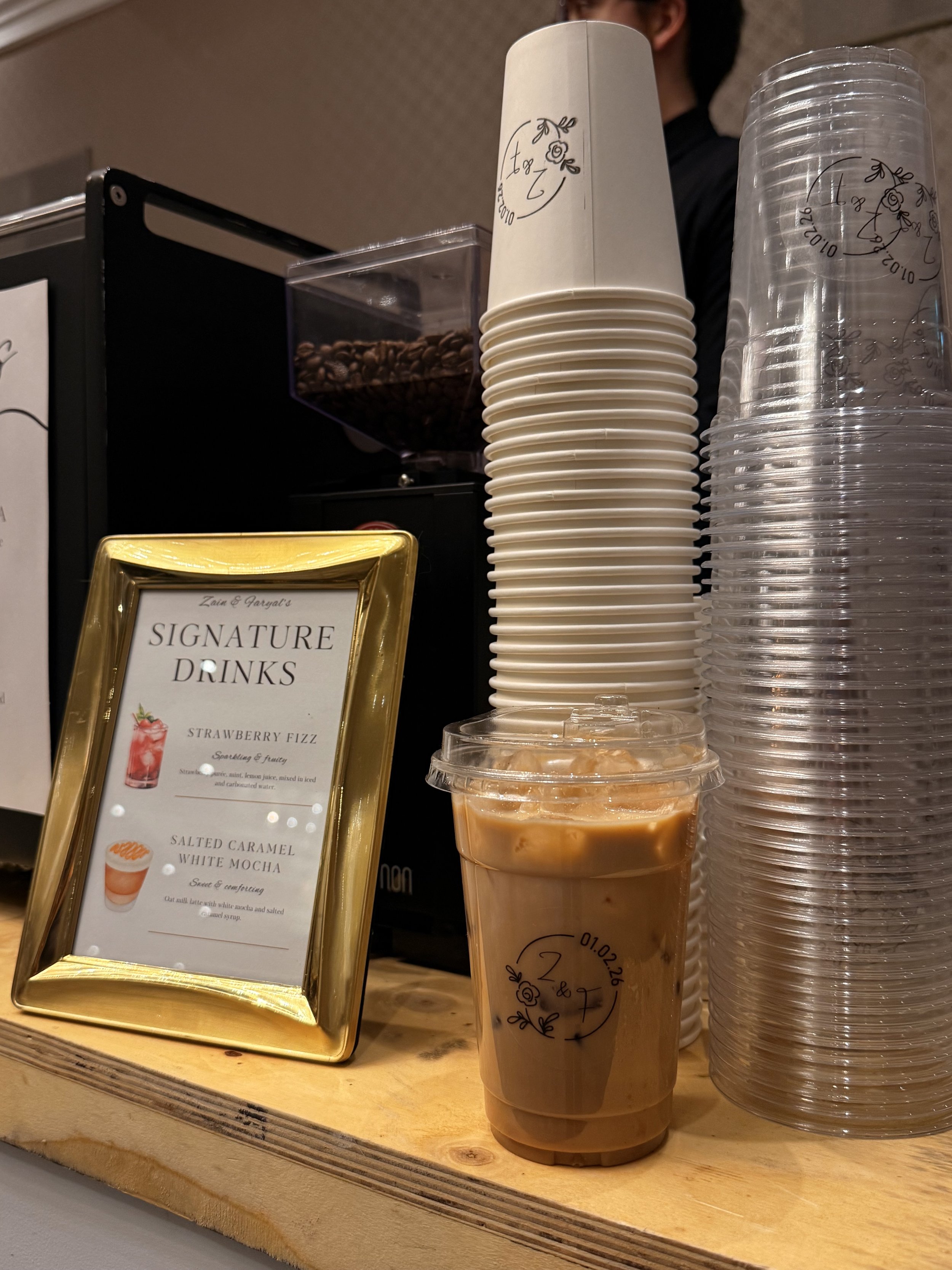 A specialty iced coffee on a wooden bar at a brand activation in Ottawa, Ontario. Stacks of hot and cold beverage cups are visible behind the drink, highlighting the variety of coffee and matcha being served.