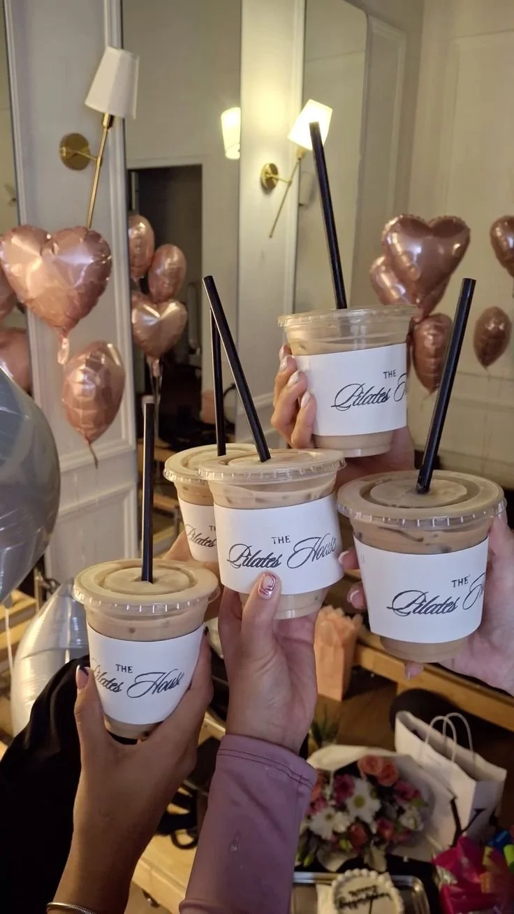 Close-up shot of four hands holding various iced and hot coffee drinks. Each cup features a prominent Pilates logo. The hands are positioned in the center of the frame as if clinking glasses together.