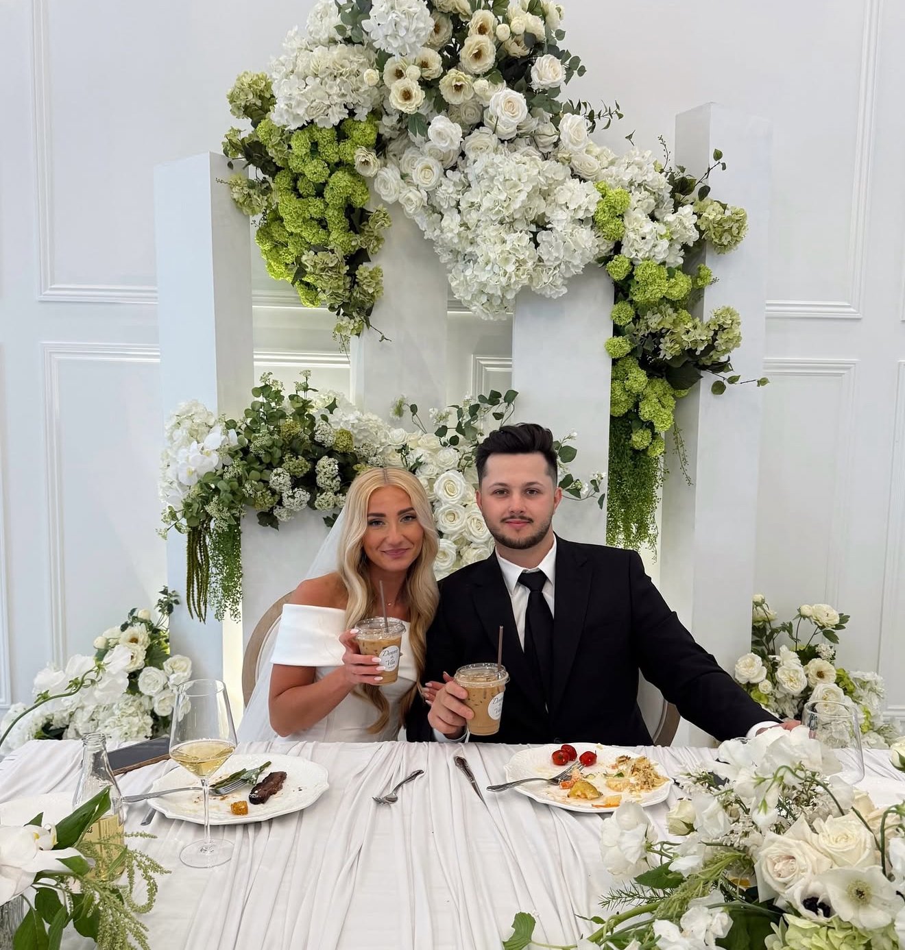 A newlywed couple sitting at a wedding reception table decorated with white flowers in Ottawa, holding signature handmade iced lattes made my the turn-key mobile espresso service made by Cafe Apero.