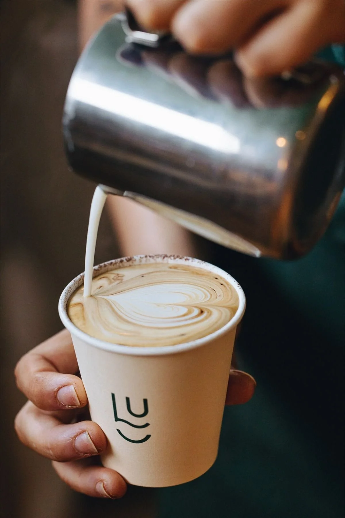 Close-up of a Café Apéro barista pouring cream into a specialty latte with intricate latte art. A hand with purple nails holds the custom-branded paper cup, showcasing the premium coffee catering experience for Montreal and Ottawa and Gatineauevents.