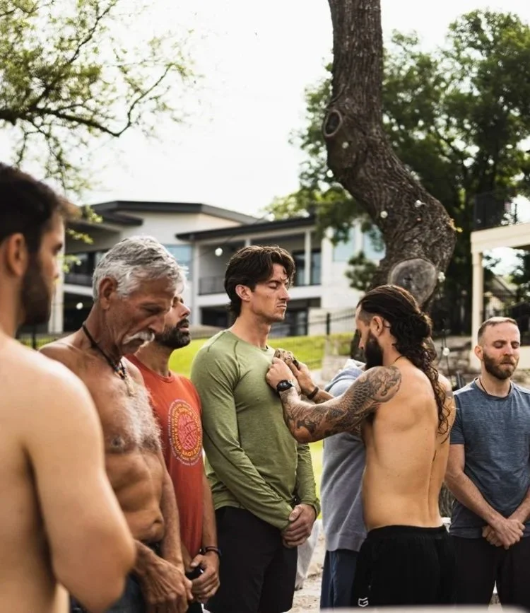 Group of men standing outdoors during a men's retreat spiritual meditation ceremony, with one shirtless man performing a ritual or blessing on another man.