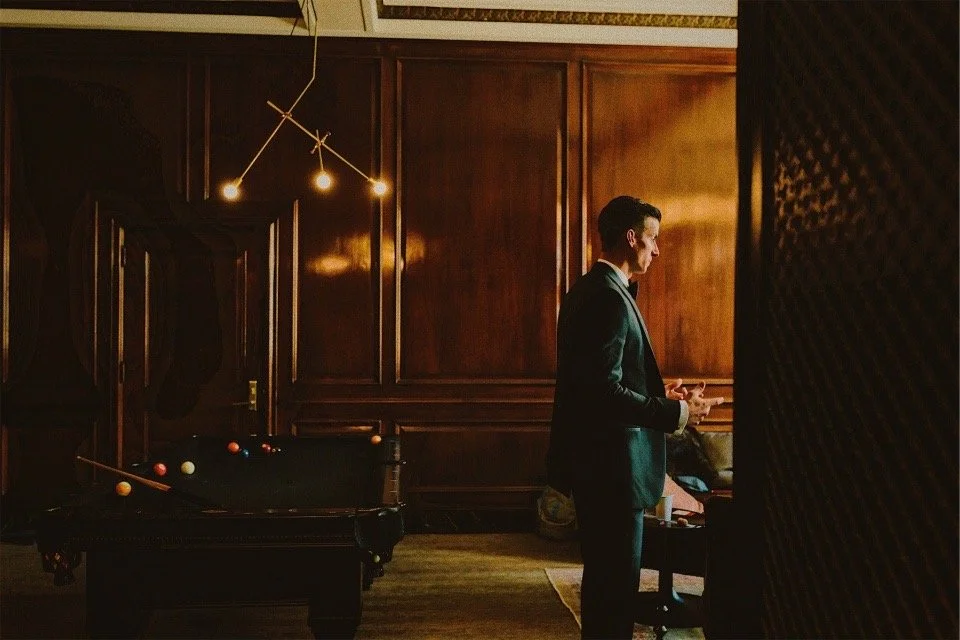 A business man in a dark suit stands in a dimly lit, wood-paneled room, with a modern gold chandelier hanging from the ceiling. Dark and calm surrounds him.