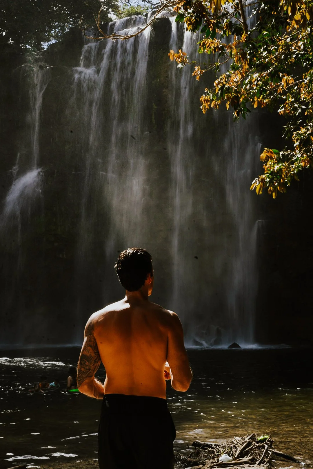 A shirtless man with dark hair and a tattoo on his left arm stands with his back to the camera, facing a waterfall in a lush forest setting in Costa Rica, pure bliss.