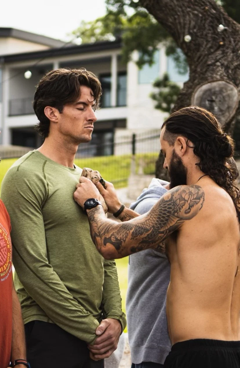 Group of men standing outdoors during a men's retreat spiritual meditation ceremony, with one shirtless man performing a ritual or blessing on another man.