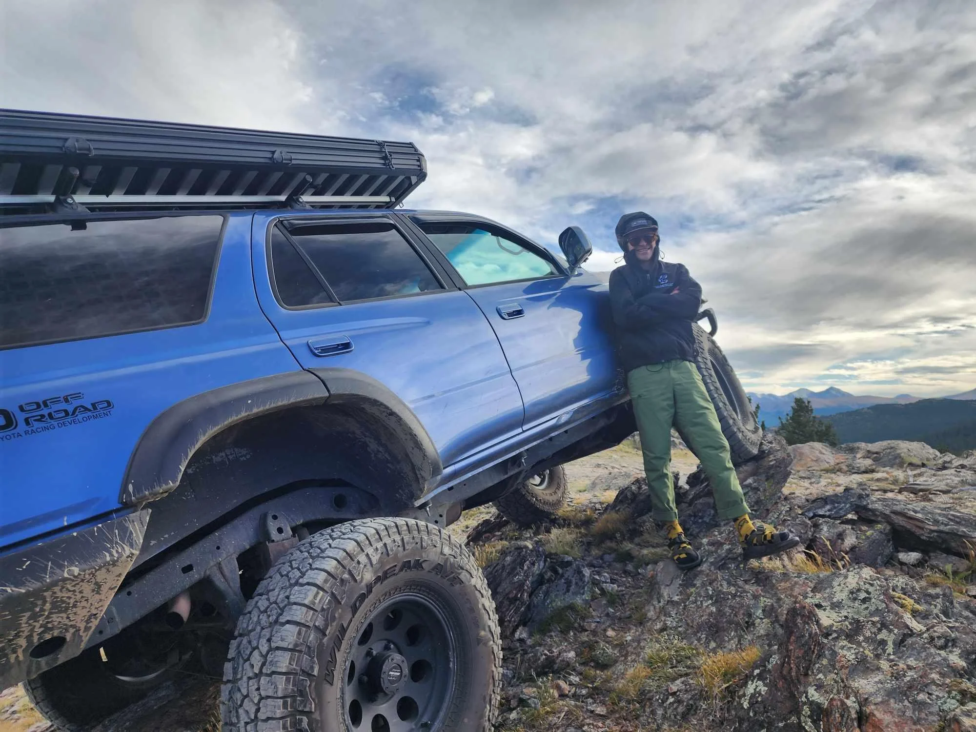 A man in outdoor gear standing beside a lifted blue vehicle on rocky terrain with mountains and cloudy sky in the background.
