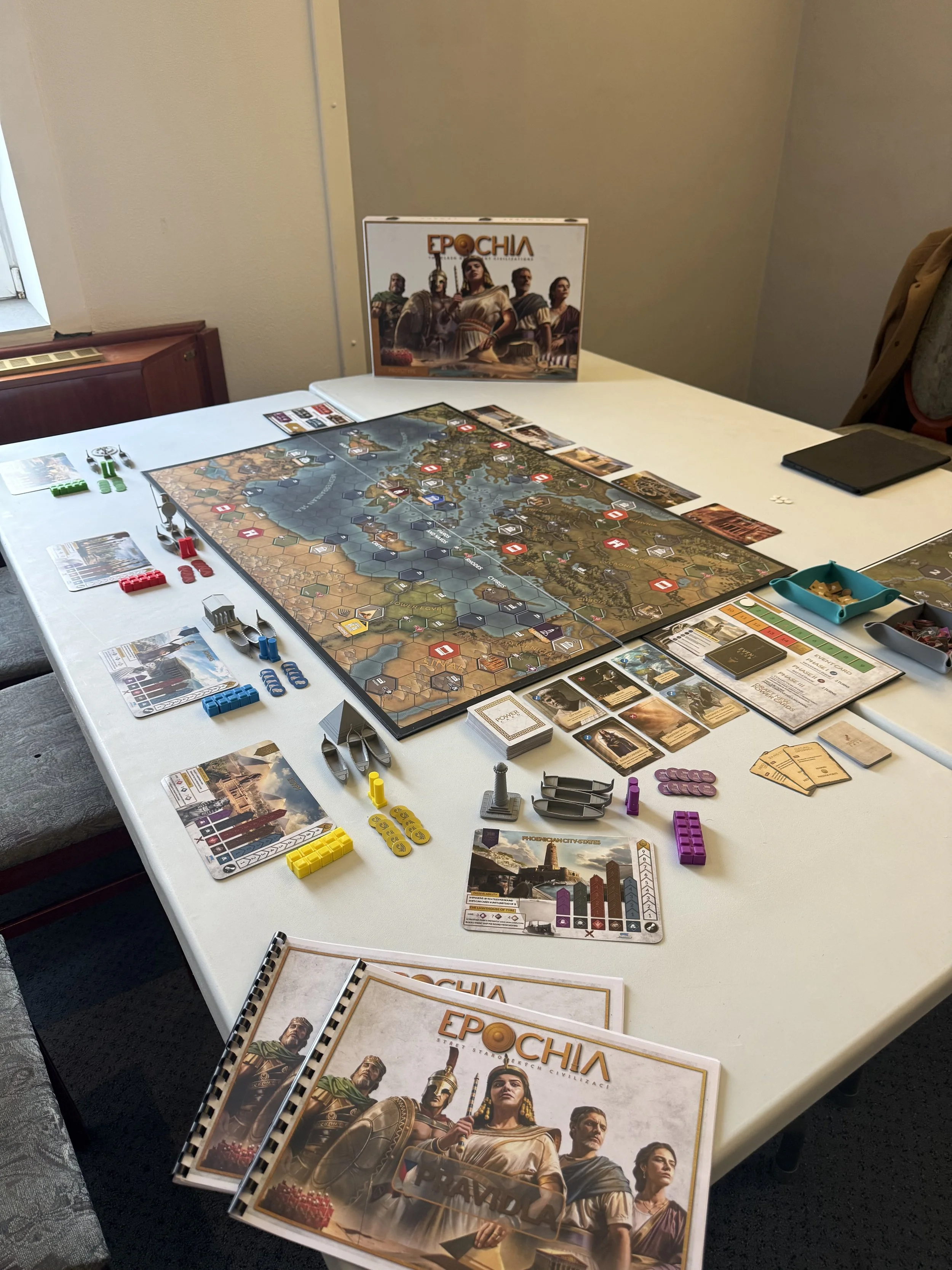 Photo of a board game setup on a white table, with a map-based game titled 'EPOCHIA' including game pieces, cards, tokens, and rulebooks, and a box of the game in the background.