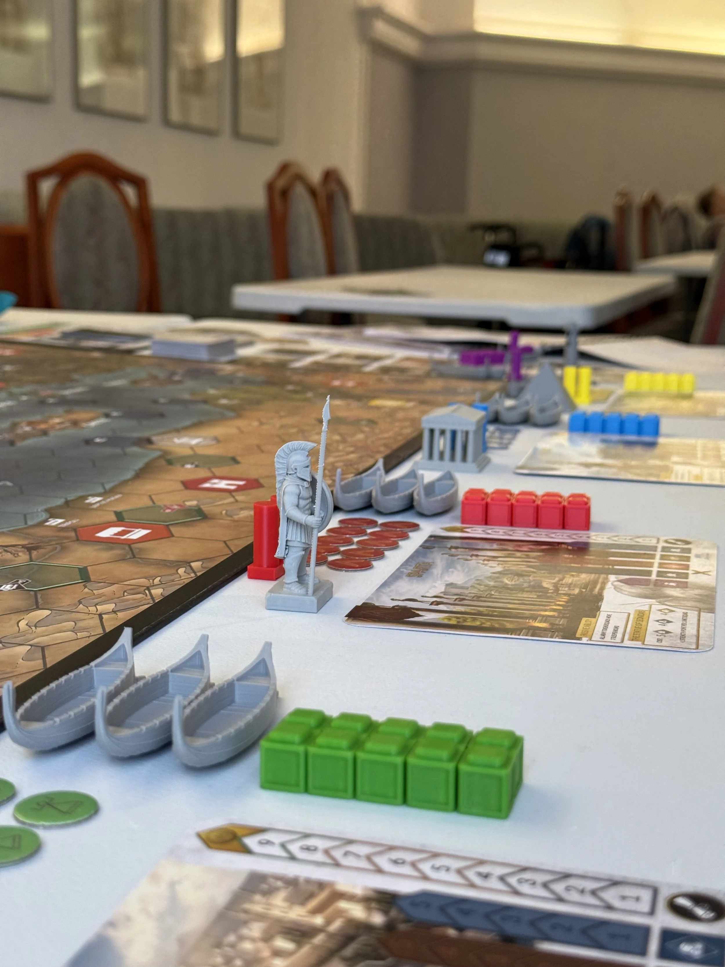Close-up of a tabletop board game with plastic figures, colored tiles, and game cards in a room with chairs and framed pictures on the wall.