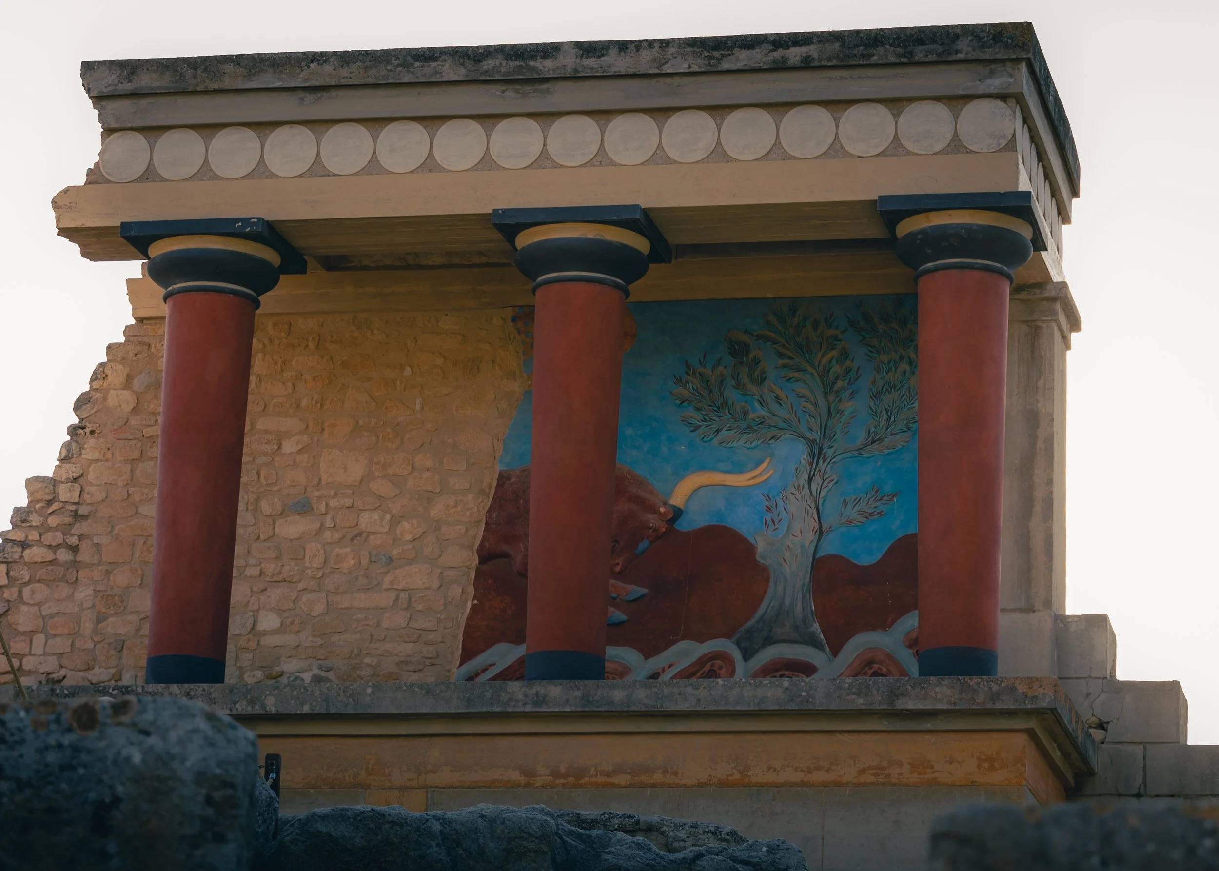 Ancient Greek-style building with three large red columns supporting a decorated entablature, featuring a painted mural of a tree and a bull on the wall behind.