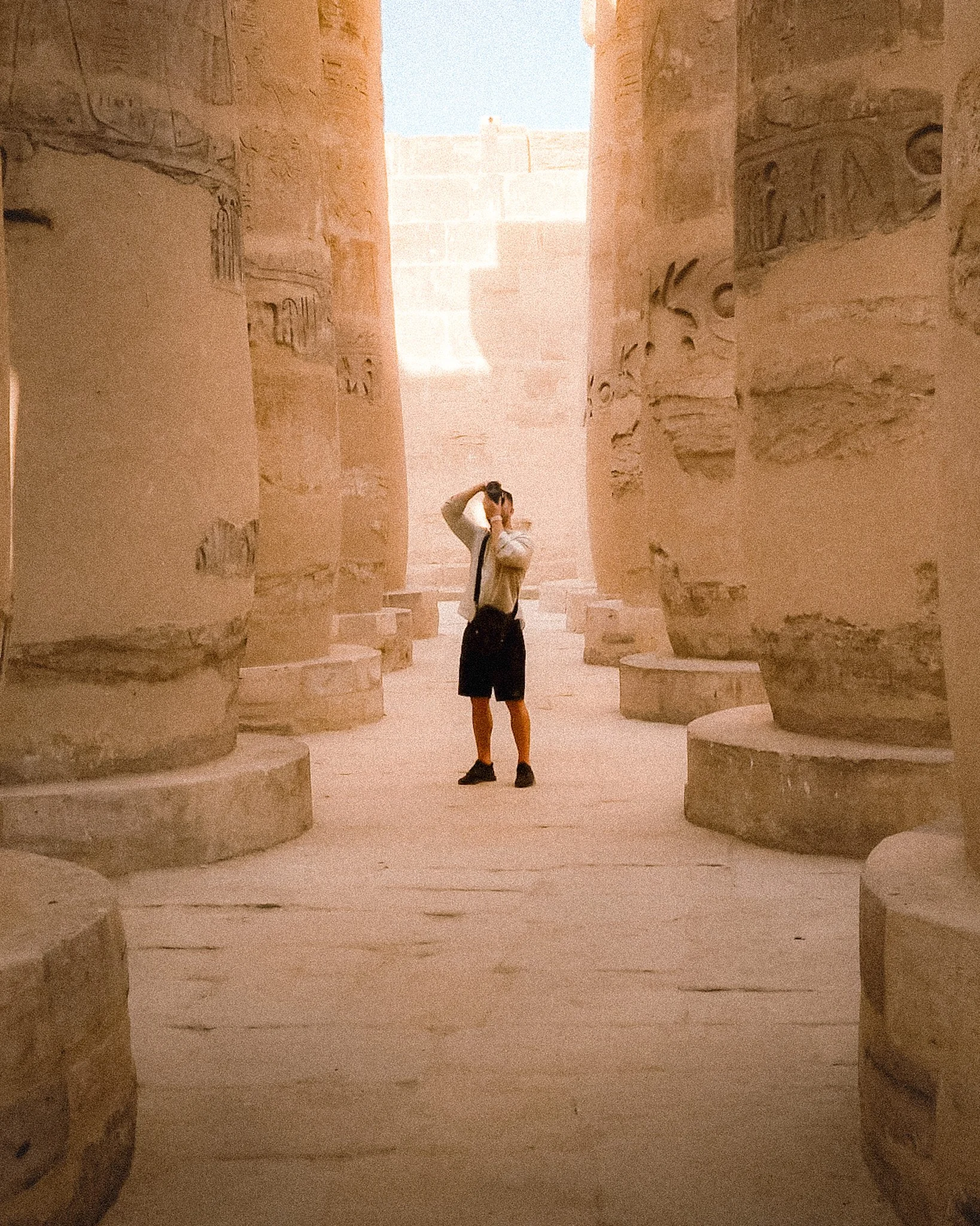 A woman standing between ancient stone walls with hieroglyphic carvings, possibly in an Egyptian temple or archaeological site, looking up and touching her head.