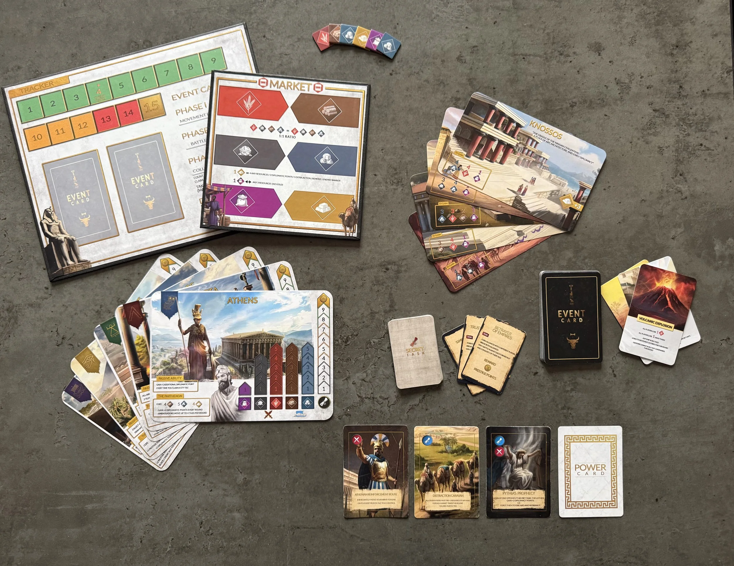 A tabletop board game setup with various cards, game pieces, and player boards. There are two decks of cards, a set of small tiles, and tokens on a grey surface.