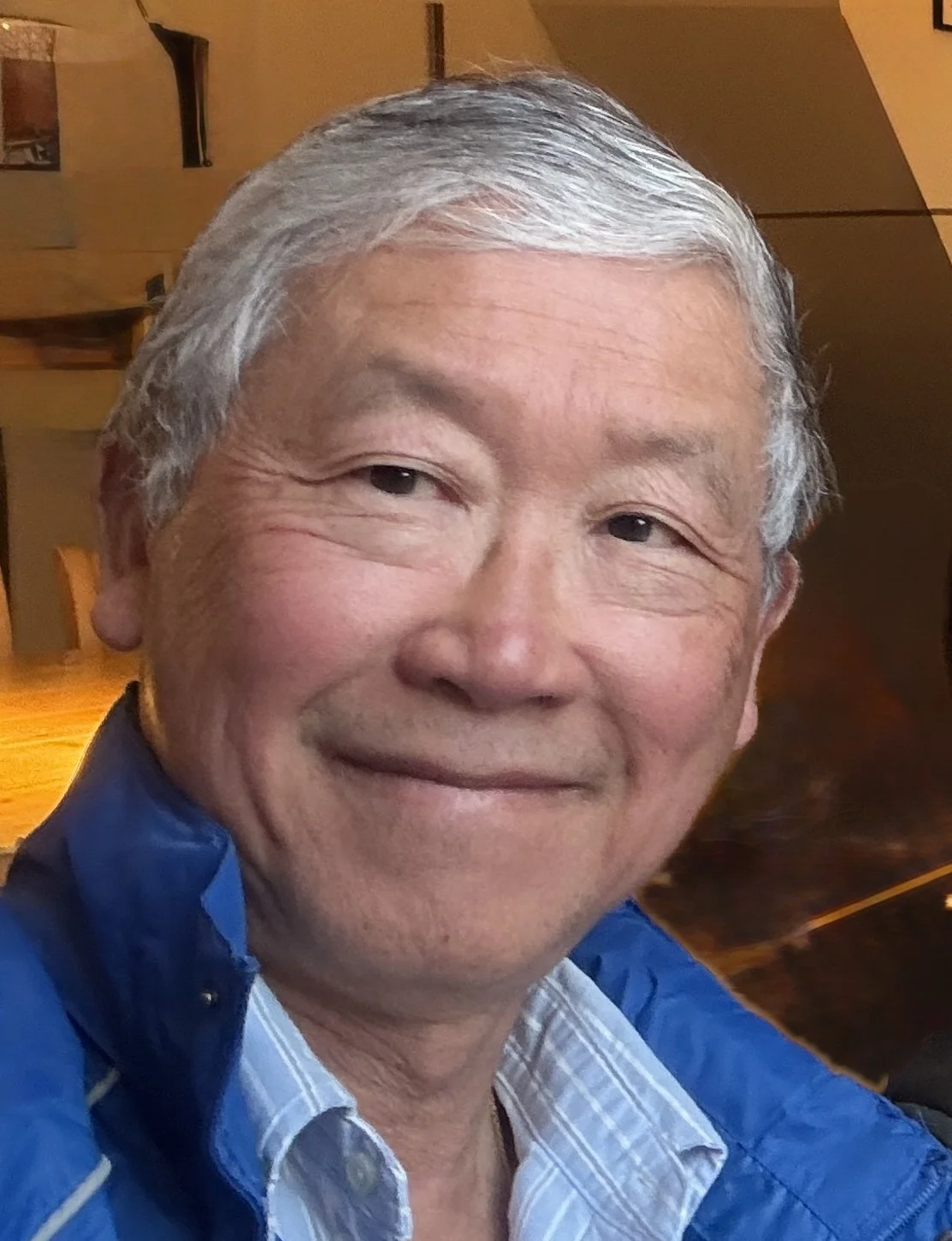 A close-up photo of an elderly Asian man with short gray hair, smiling, wearing a blue jacket over a collared shirt, in a warmly lit indoor setting.