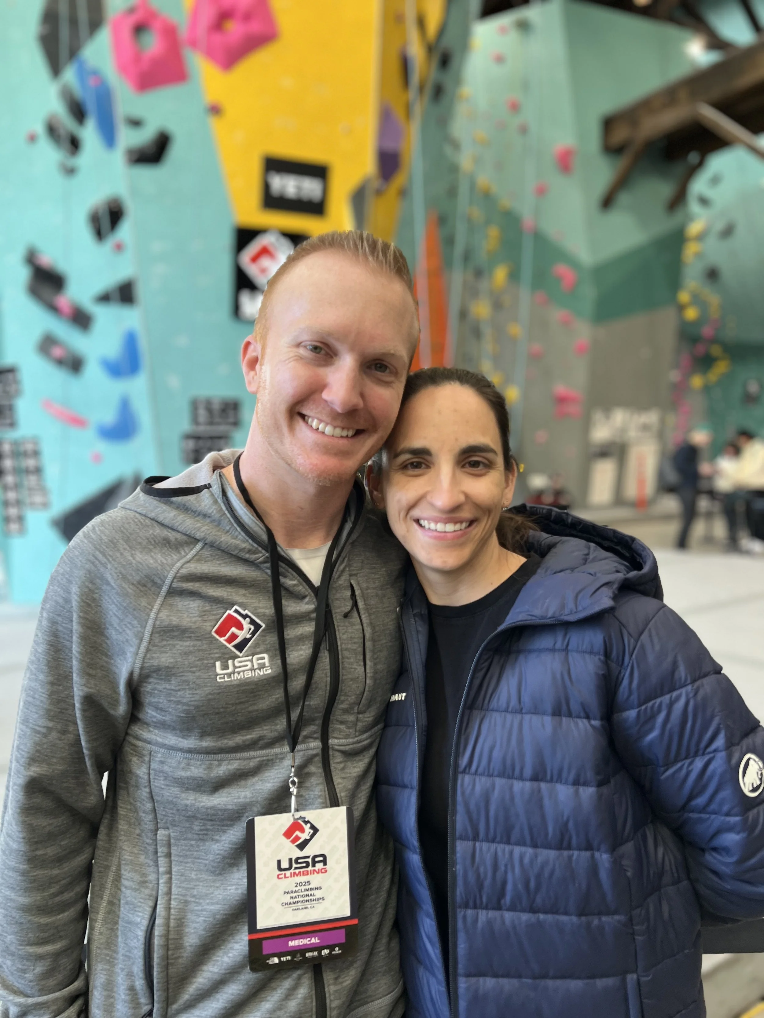 Two smiling people standing close together inside a climbing gym with colorful climbing walls in the background, one wearing a gray USA climbing jacket and the other in a blue jacket.