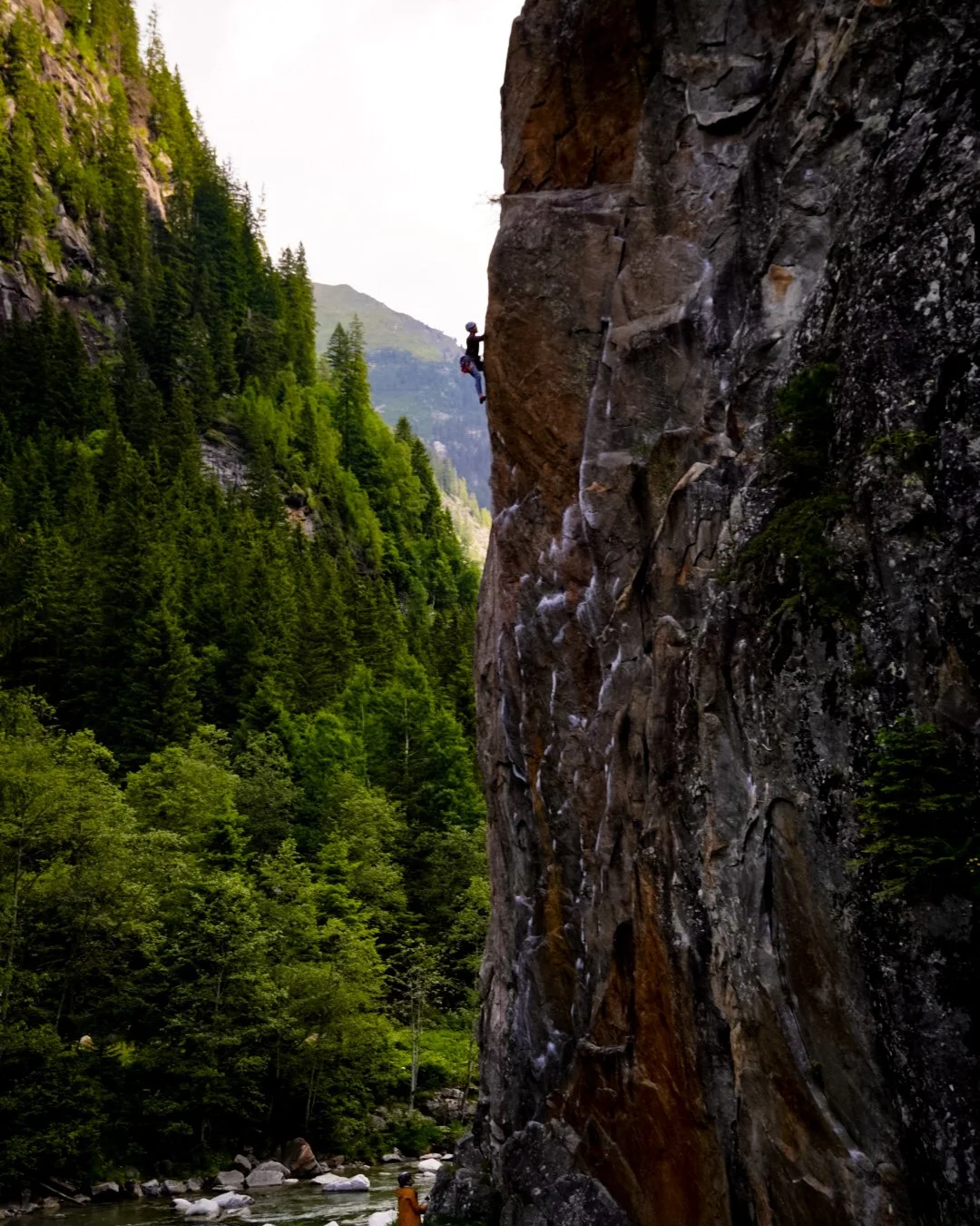 A person is rock climbing on a tall cliff face in a forested mountain area, with dense green trees and a river below.
