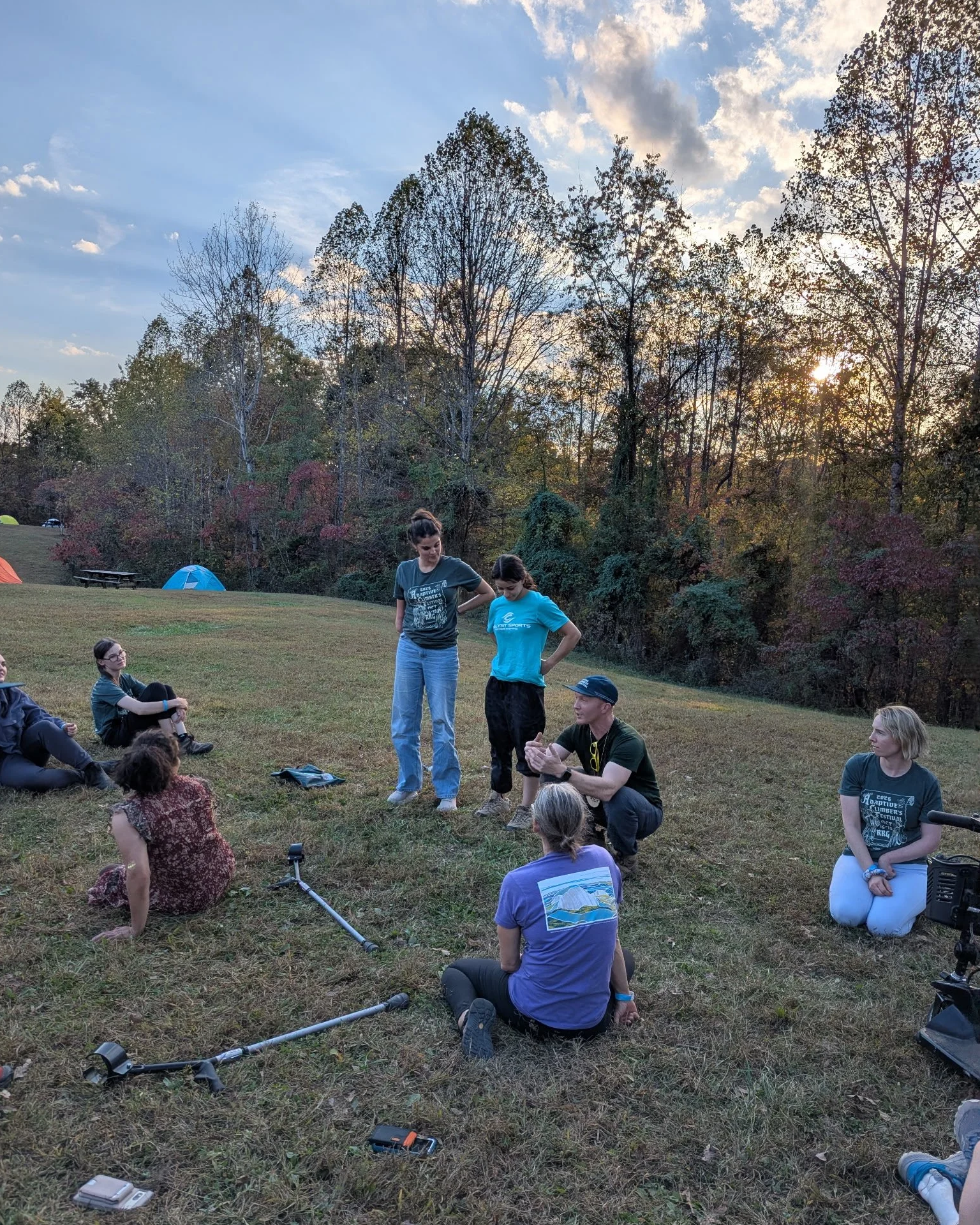 Group of people outdoors on a grassy field, some sitting and some standing, with tents in the background and trees at sunset.