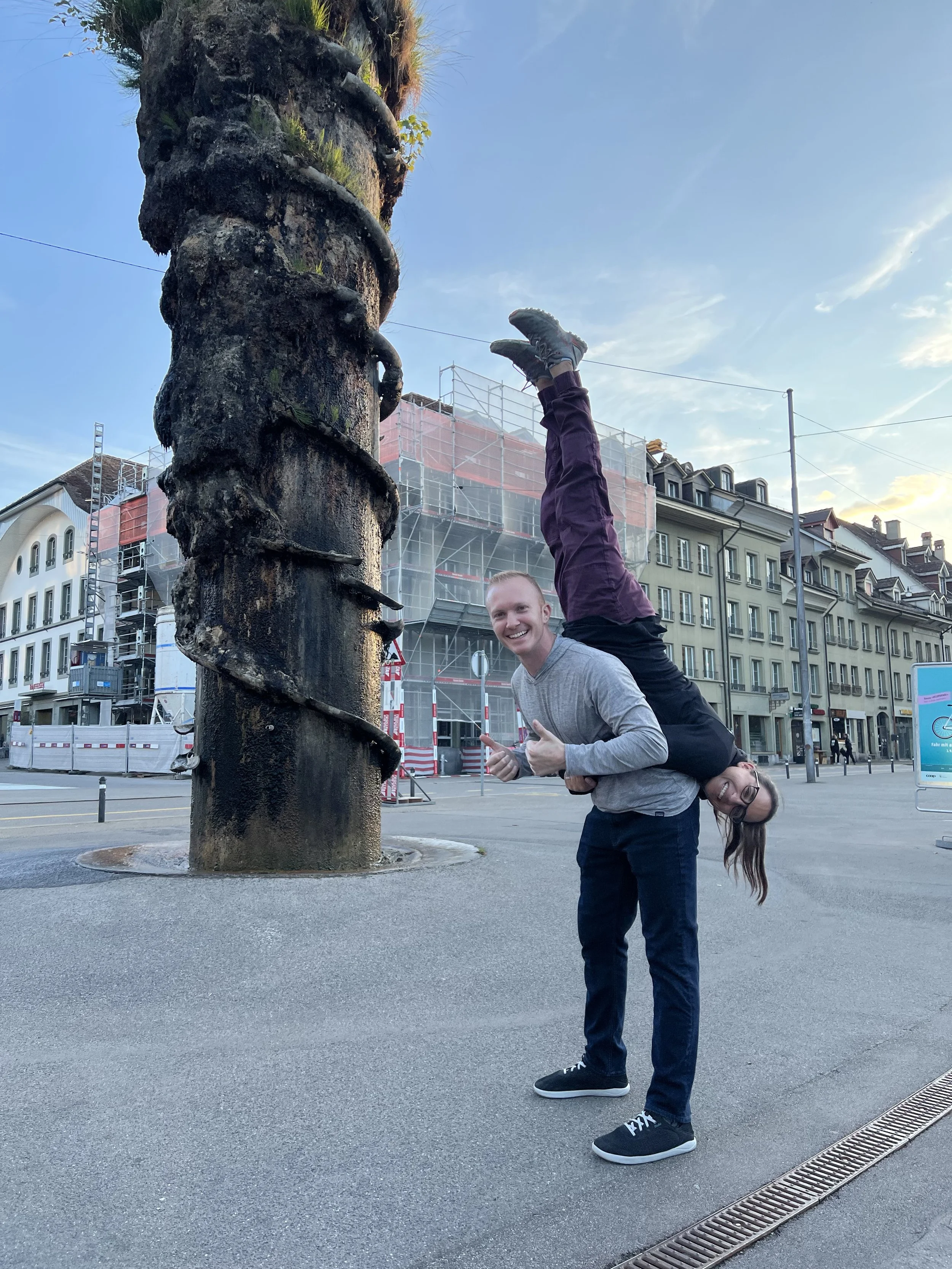 Two people performing an acrobatic pose on a city street with buildings in the background. One person is standing and supporting the other person, who is upside down with legs extended upward and head near the ground. Both are smiling.