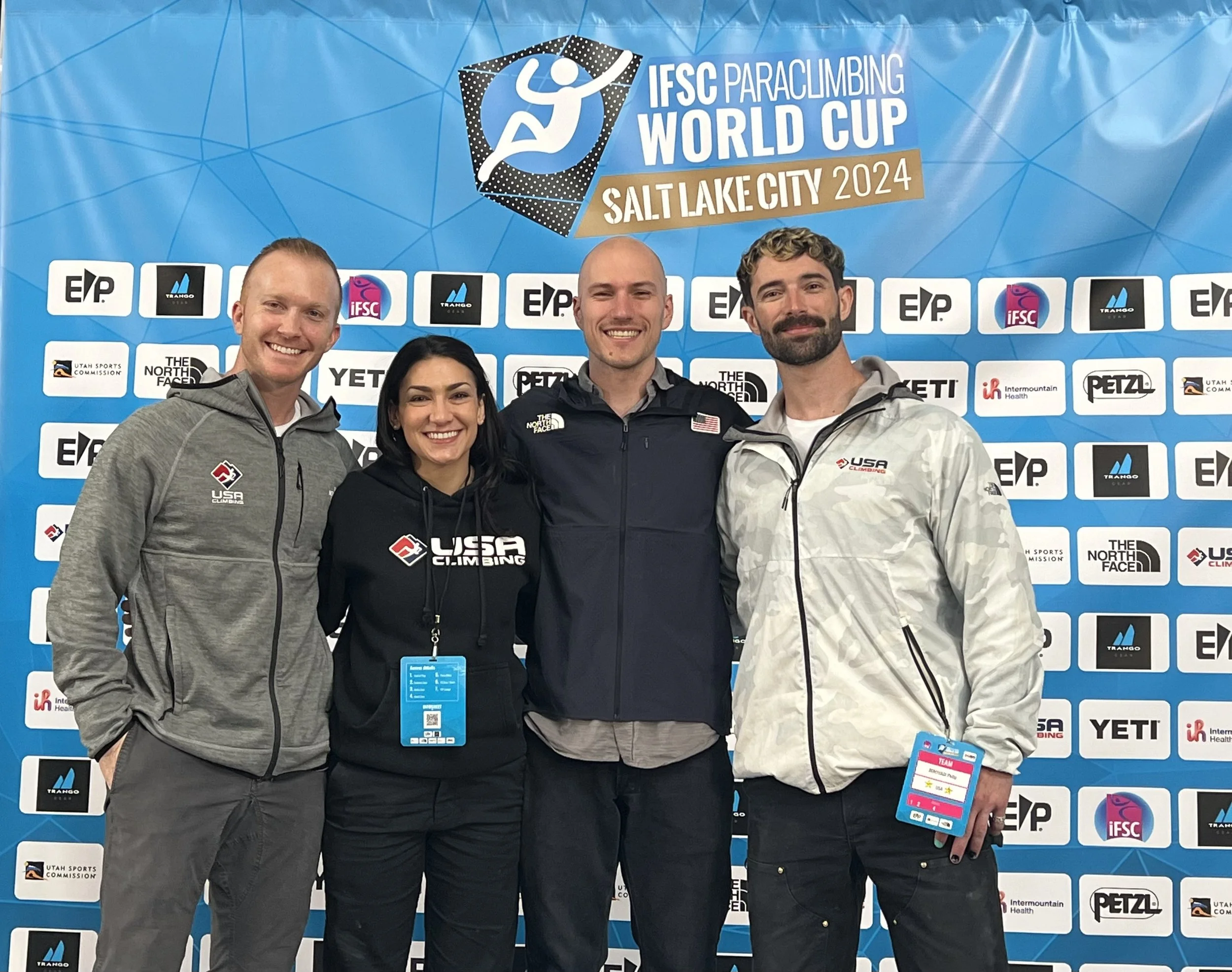 Four athletes smiling and posing in front of a blue backdrop with logos, at the IFSC Paraclimbing World Cup Salt Lake City 2024, with some wearing USA climbing jackets and badges.
