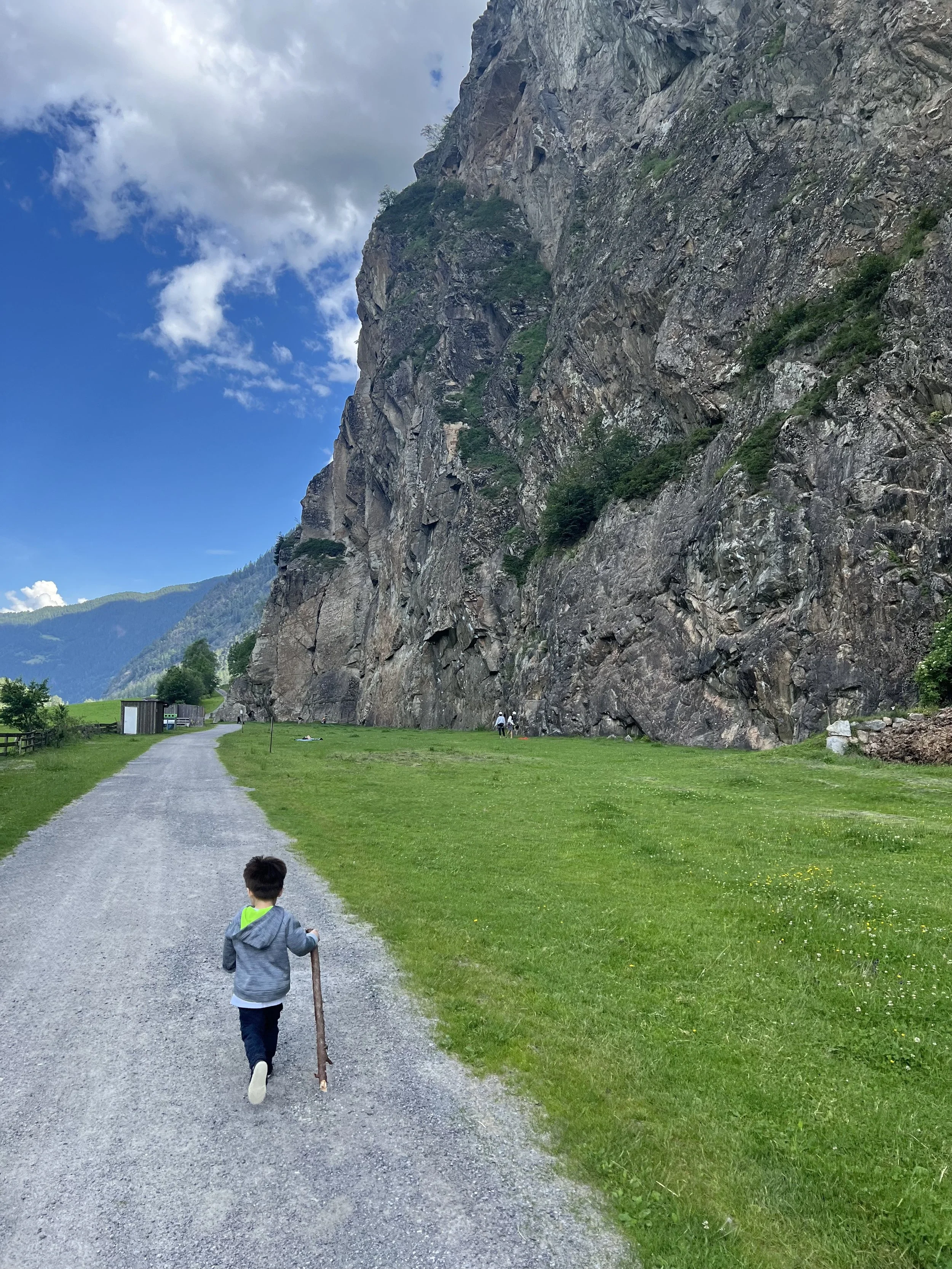 Child walking on a gravel path with a stick in hand, heading towards a large rocky cliff on a bright day with partly cloudy sky.
