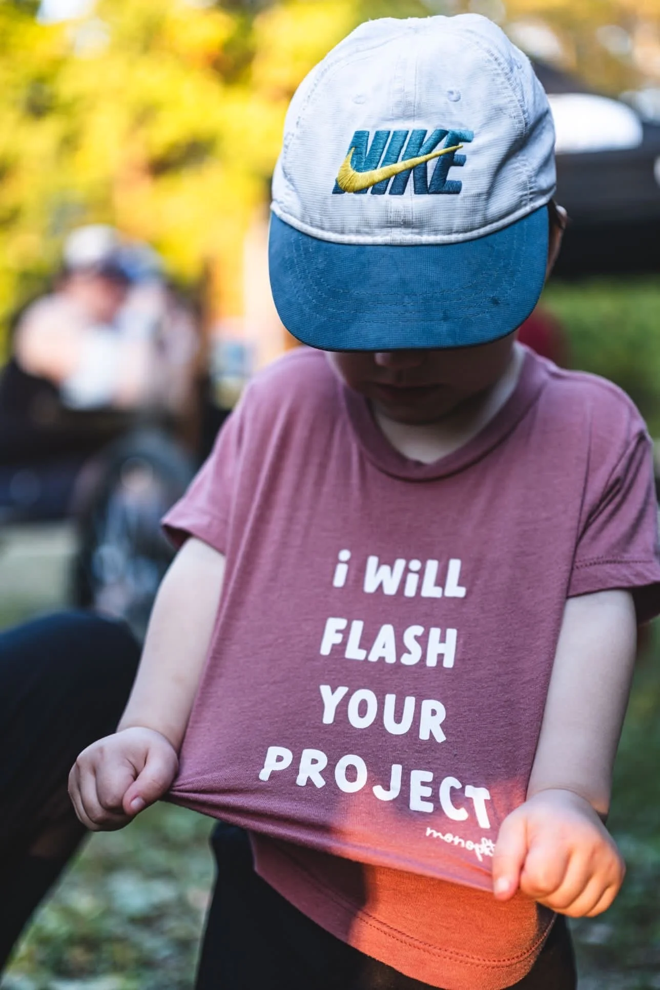 Child wearing a pink t-shirt with the text 'I WILL FLASH YOUR PROJECT' and a blue and white Nike cap, outdoors with blurred background.