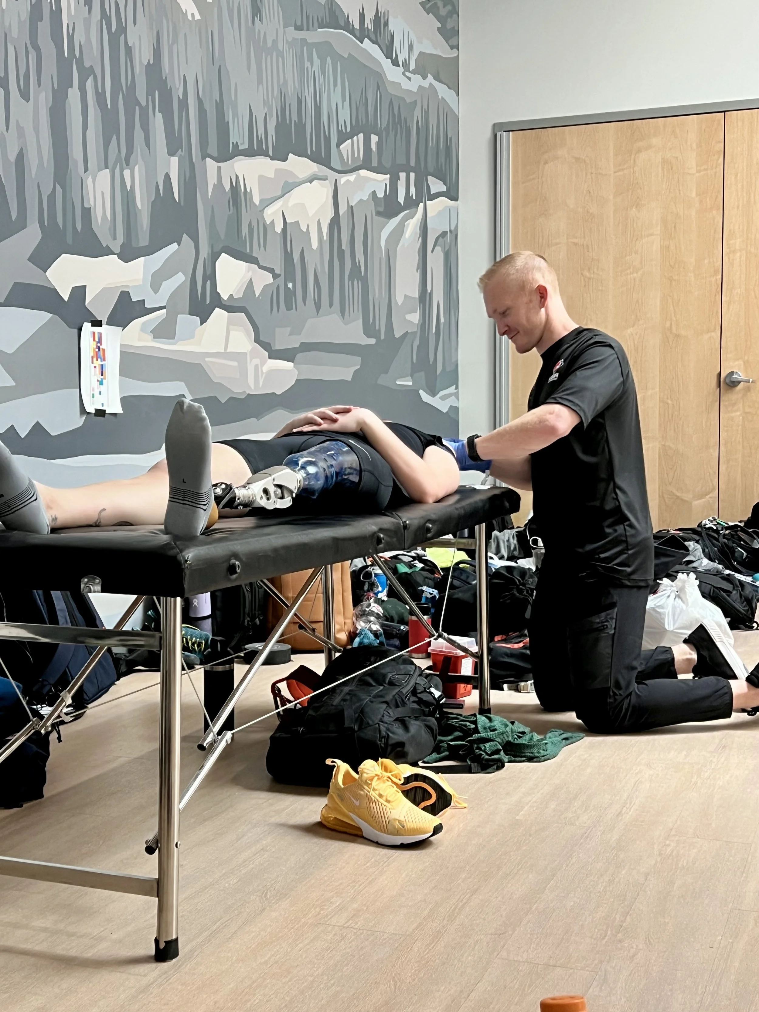 A person with a prosthetic leg receiving treatment from a medical professional on a massage table, surrounded by sports gear and bags in a room with a mountain mural on the wall.